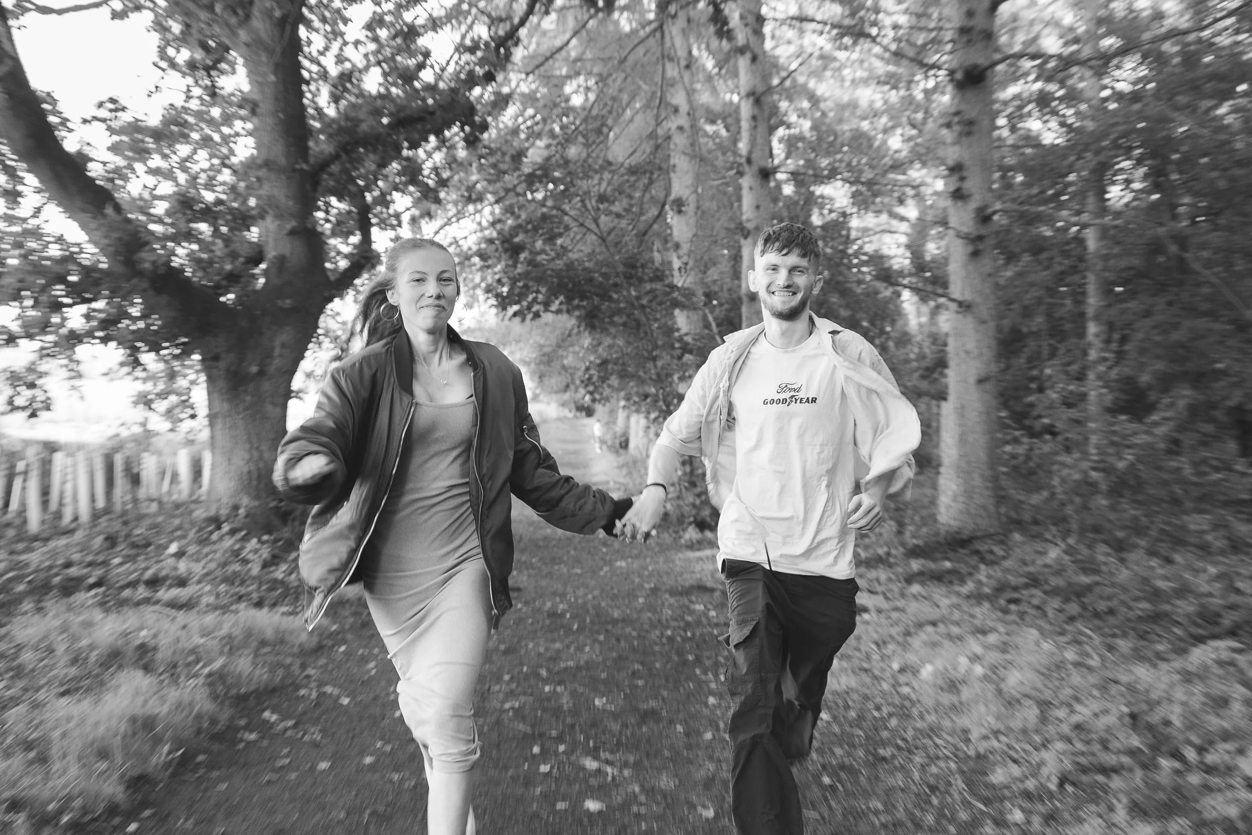 A black and white photo of a young woman and man running outdoors along a tree-lined path, holding hands and smiling.