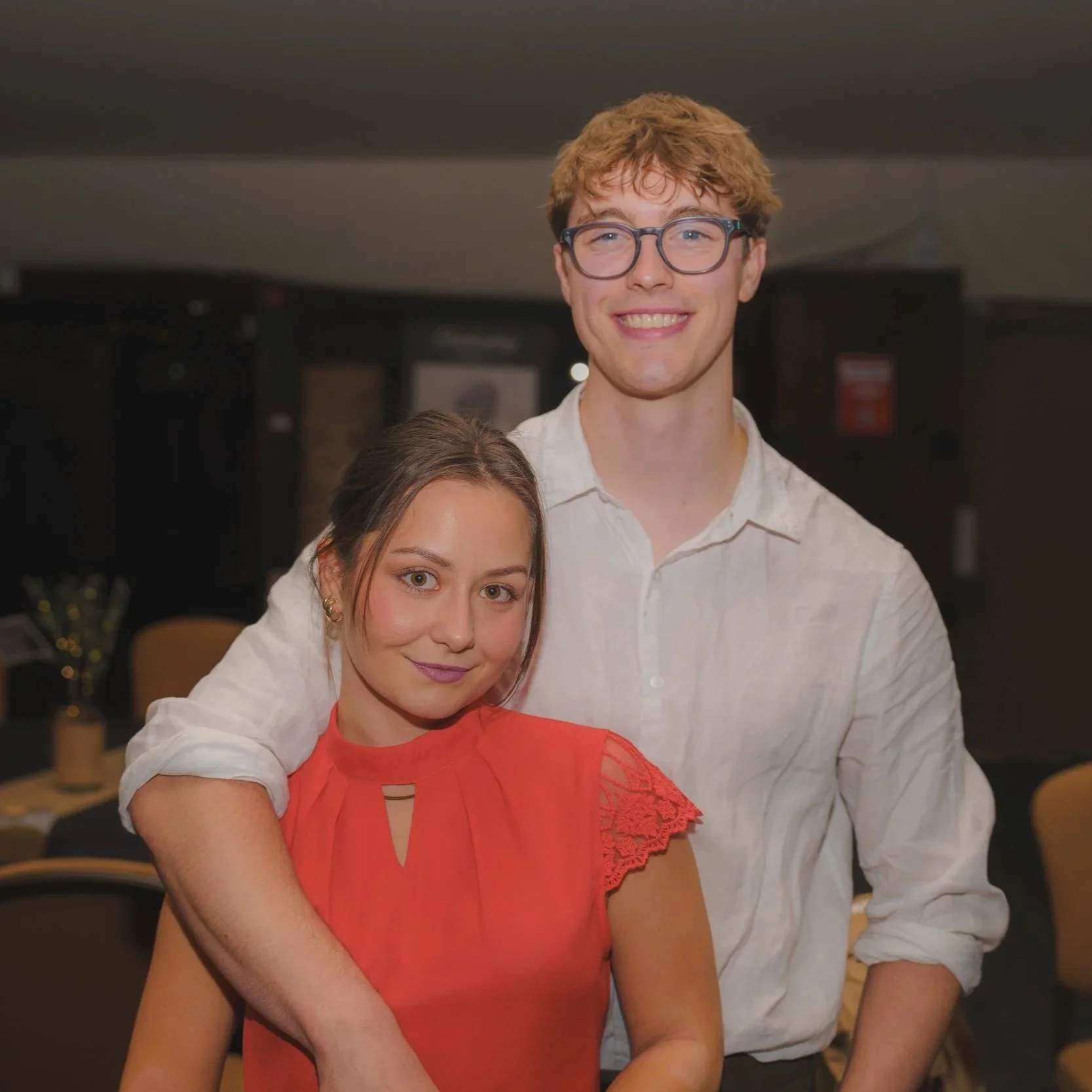 A young man with glasses and light brown curly hair smiling, standing next to a young woman with brown hair, green eyes, and wearing an orange dress with lace details.