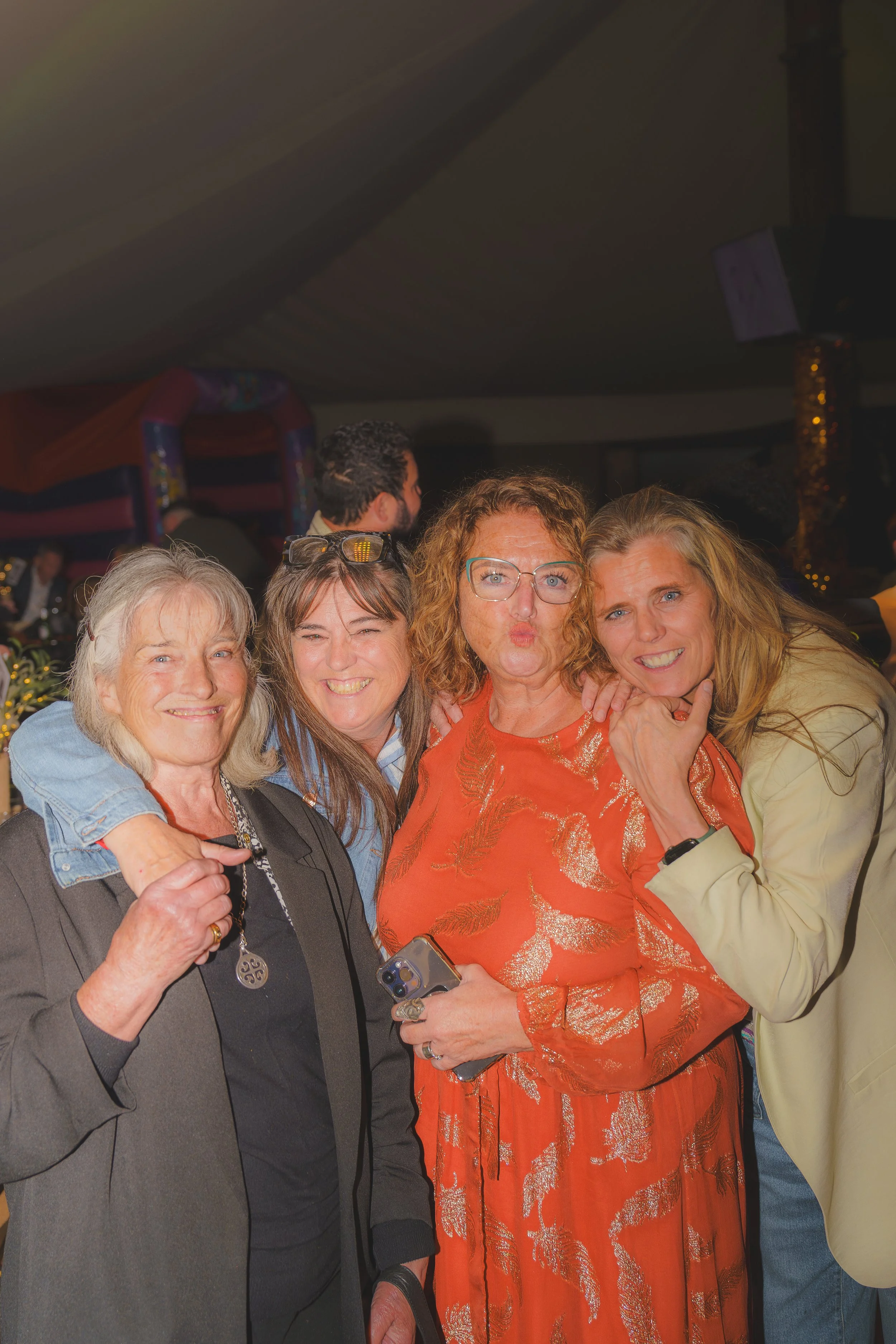 Four women posing together at an indoor event, smiling and embracing, with background showing other people and blurred decorations.