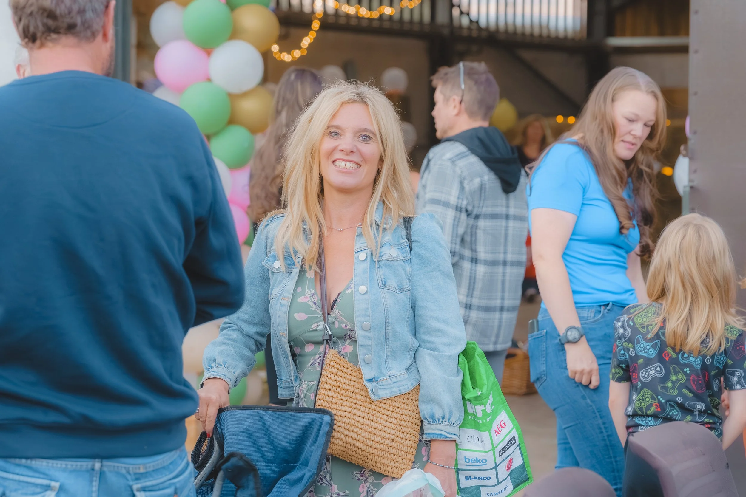 People at a social gathering, including a smiling woman with blonde hair wearing a denim jacket, surrounded by others in casual attire, colorful balloons, and festive lighting.