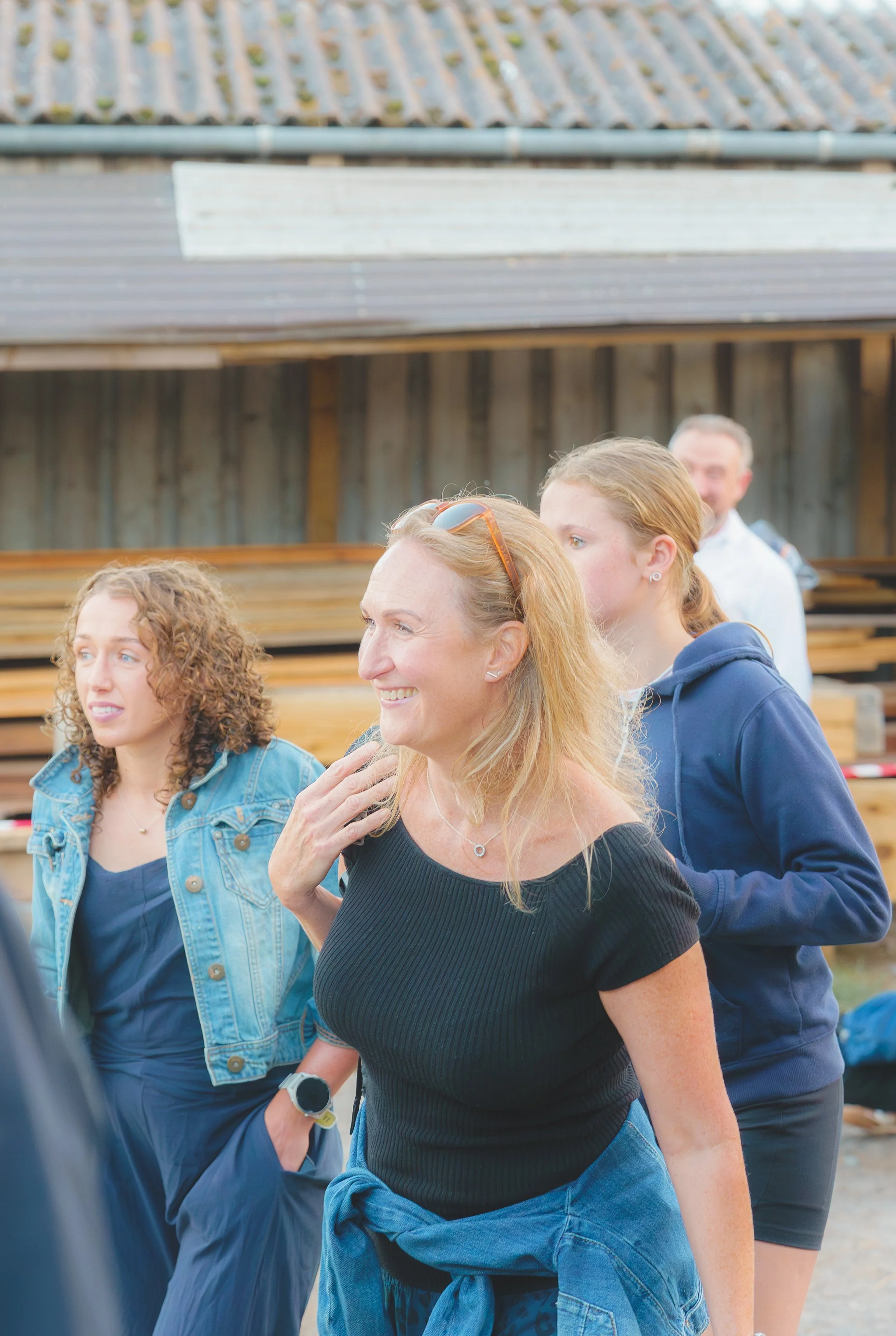 A group of people walking outdoors, with a woman in the foreground smiling, wearing a black shirt and blue jeans, and others around her including a woman with glasses and a woman with red hair. They are in front of a wooden fence and a house with a t