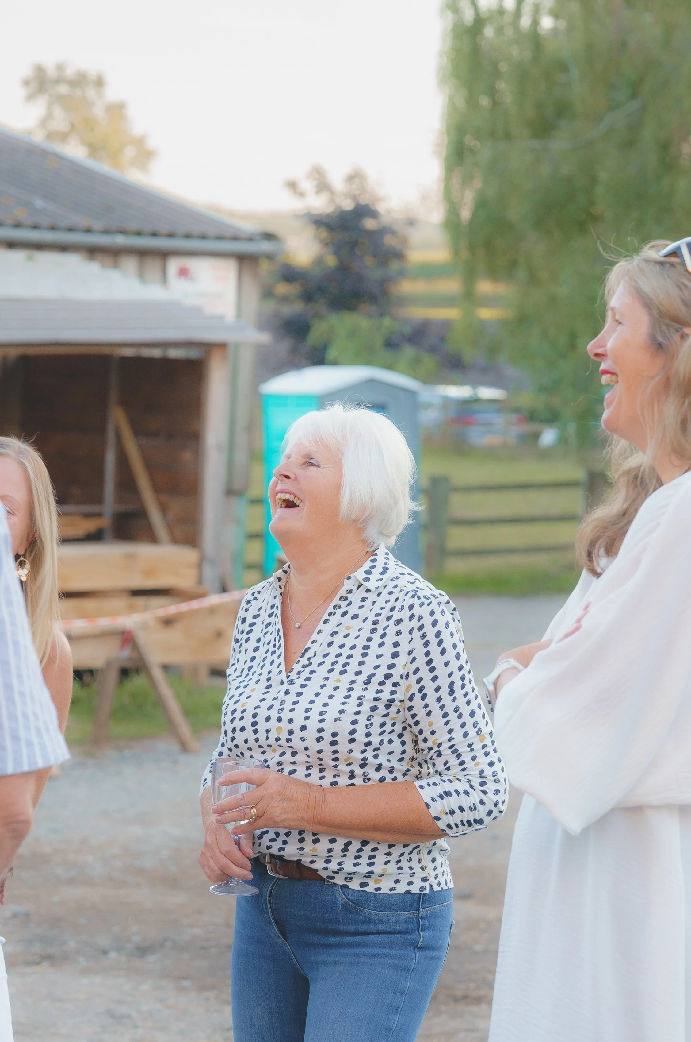 A group of women laughing and socializing outdoors during daytime, with a woman holding a wine glass in the foreground.