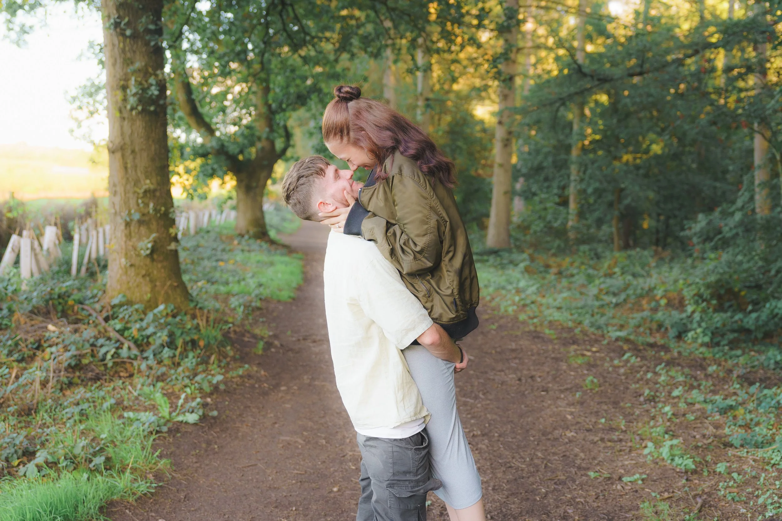 A man is holding a woman in his arms on a wooded path, and they are touching foreheads and smiling at each other.