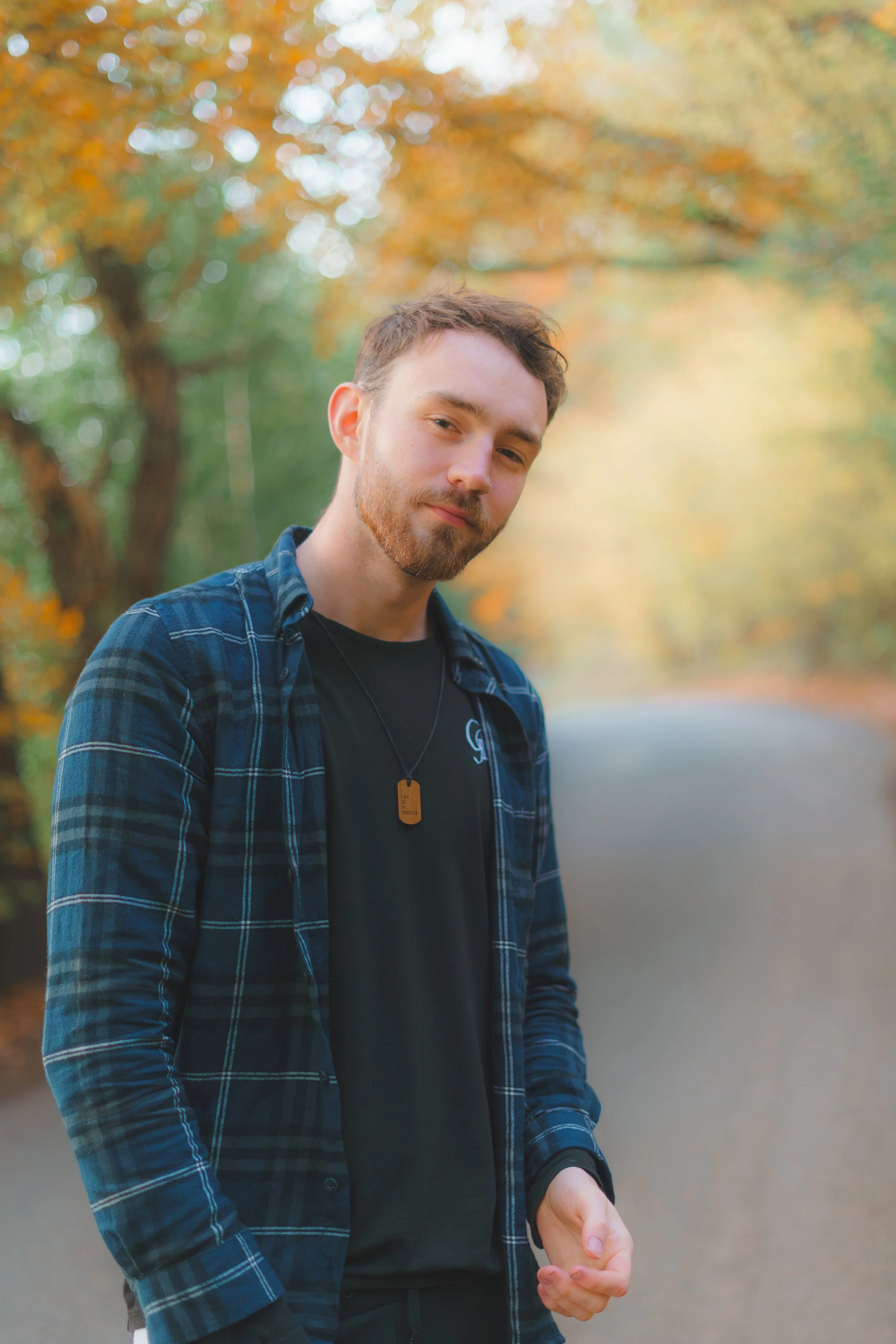 A young man with short brown hair, a beard, and light skin stands outdoors on a path surrounded by trees with fall-colored leaves. He wears a blue and black plaid shirt over a black T-shirt. The background features blurred orange, yellow, and green foliage.