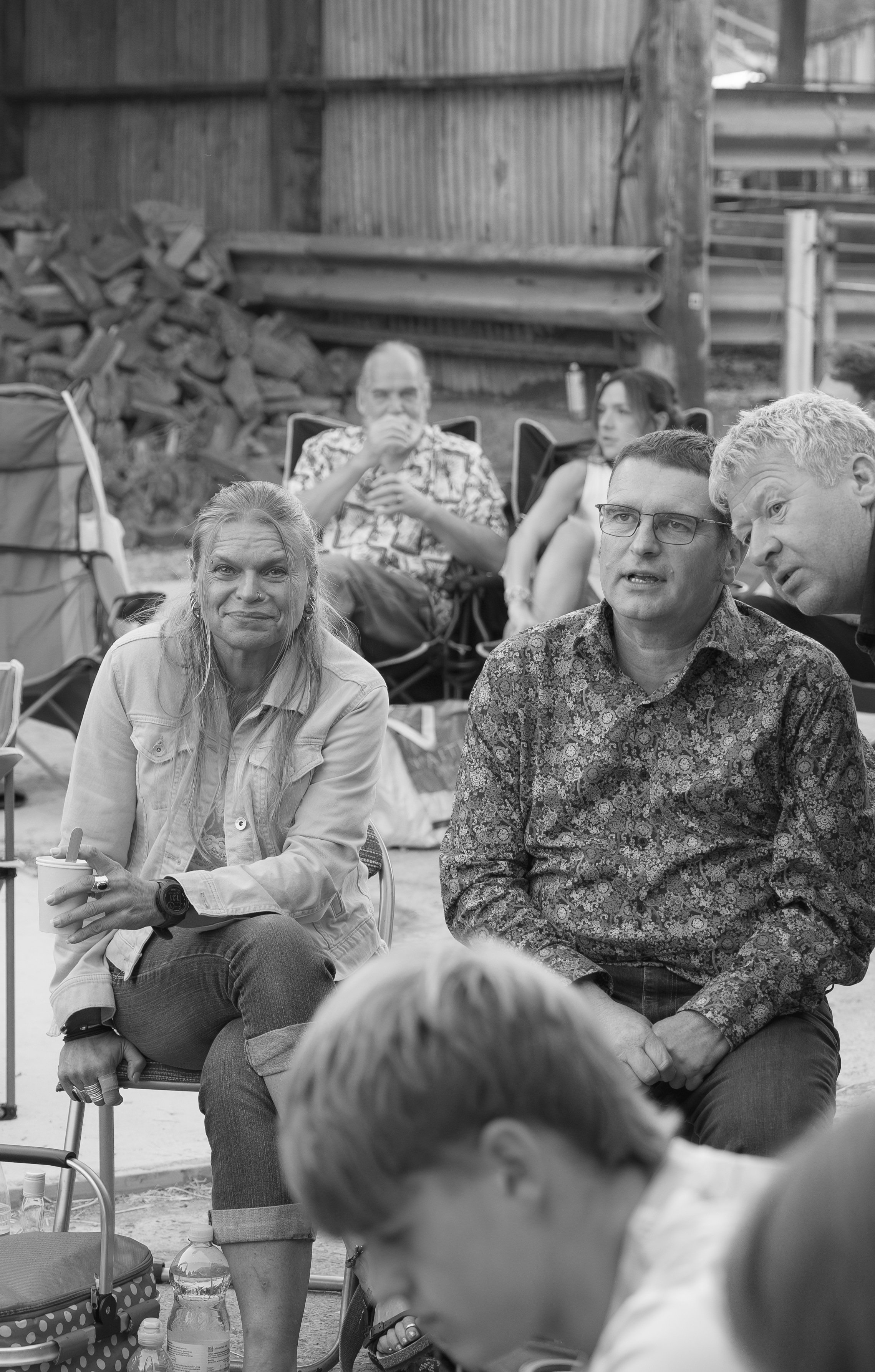A group of people sitting outdoors, engaged in conversation, with a wooden fence and stacked firewood in the background, in a black-and-white photo.