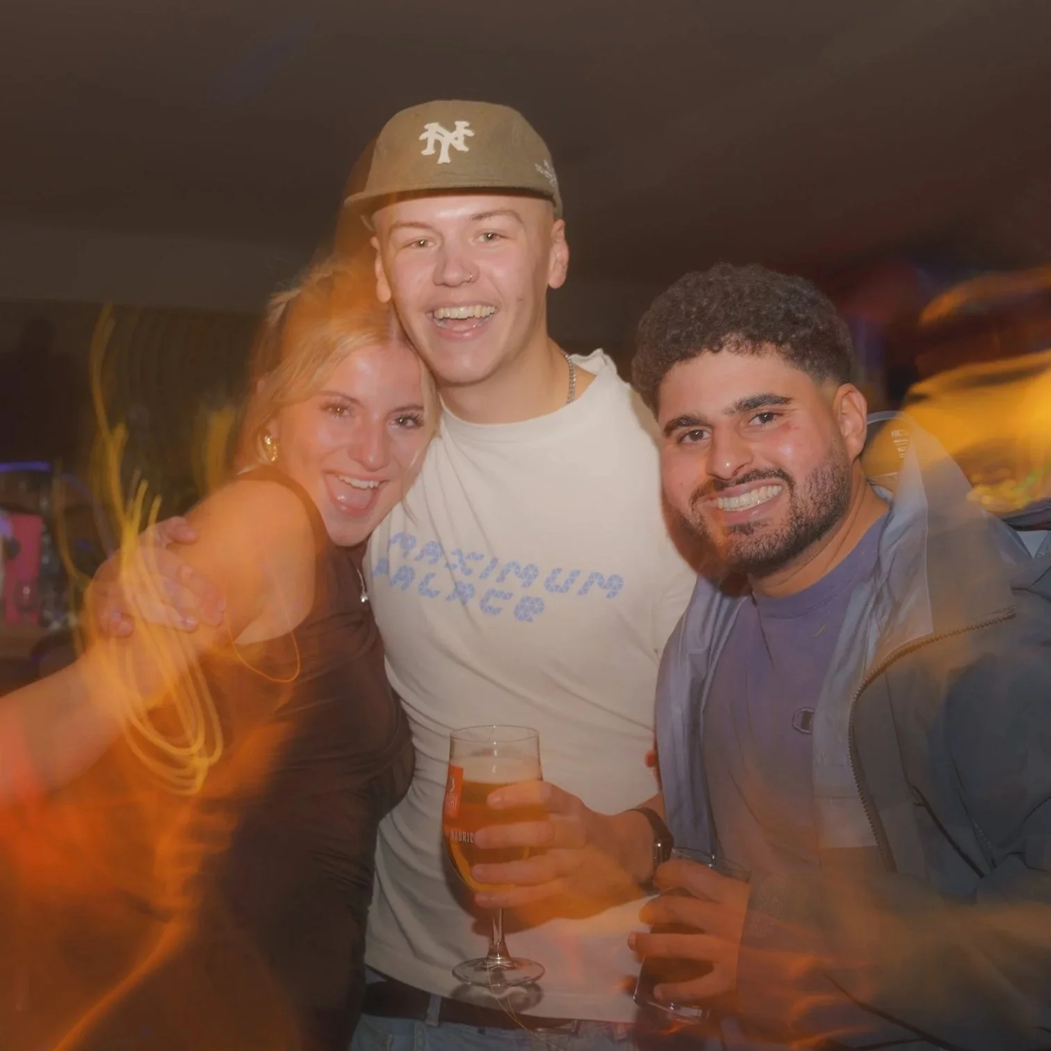 Three smiling friends at a party, holding beer glasses, with a blurred background and some light streaks.