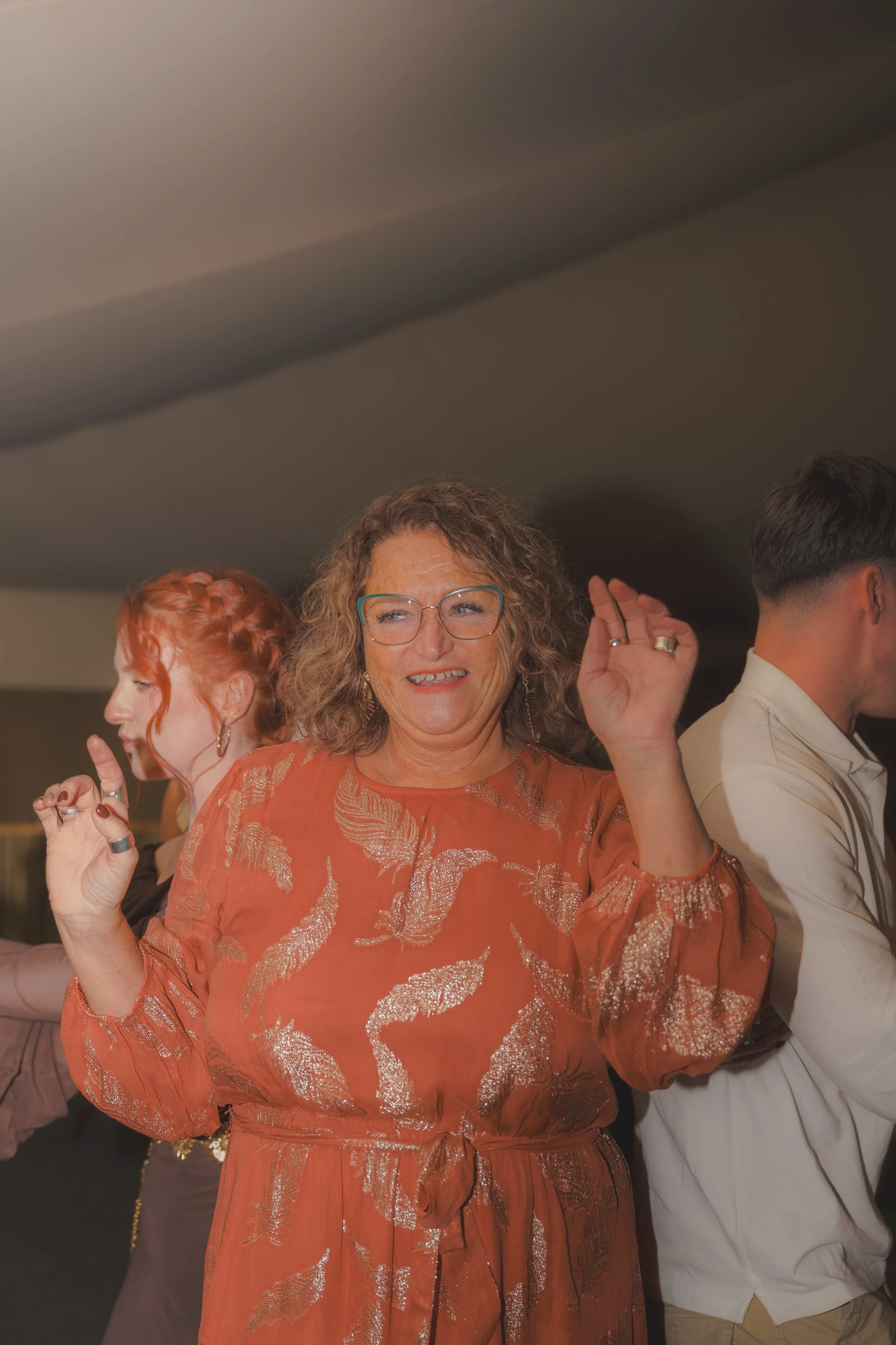 A woman with curly hair wearing glasses and an orange dress with silver leaf patterns, dancing with a smile and closed eyes. She is surrounded by other people at a social gathering or celebration.
