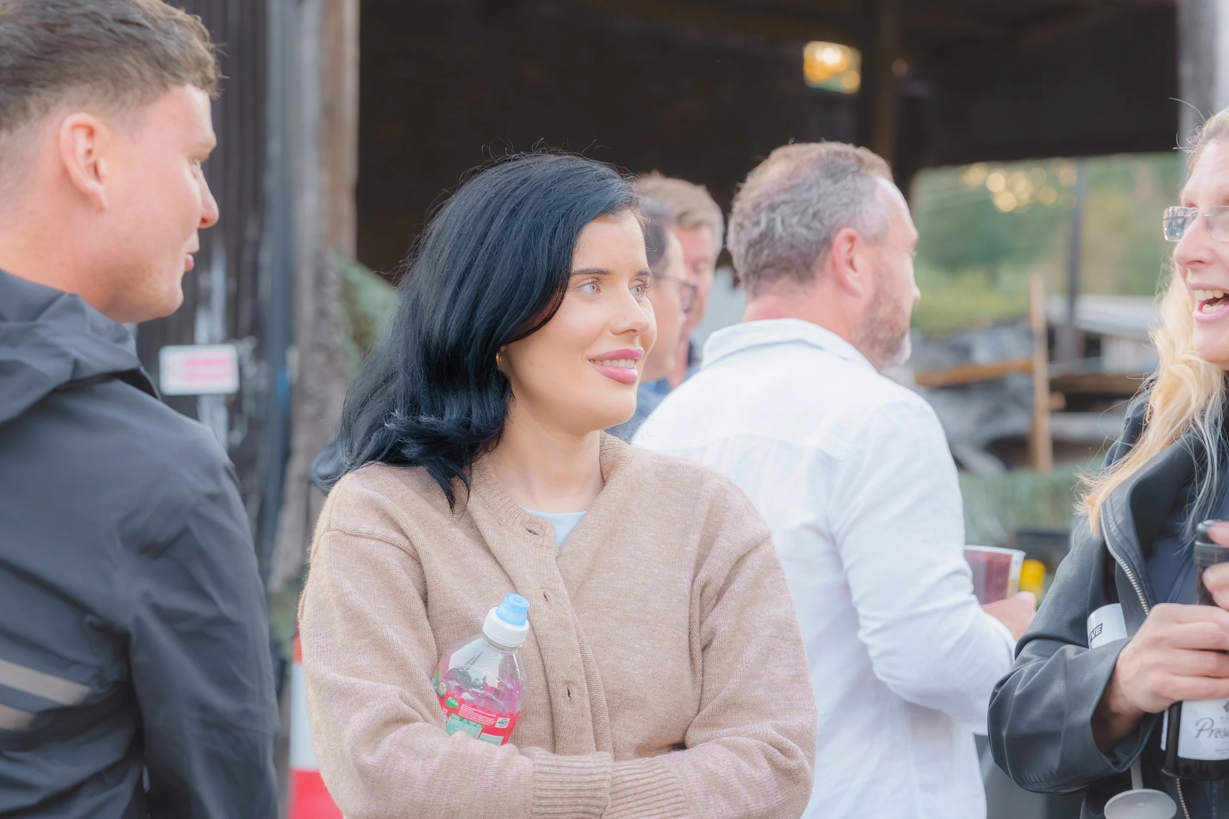 A group of people socializing outdoors, including a woman with black hair holding a water bottle, smiling, and talking to a blonde woman wearing glasses and a black jacket.