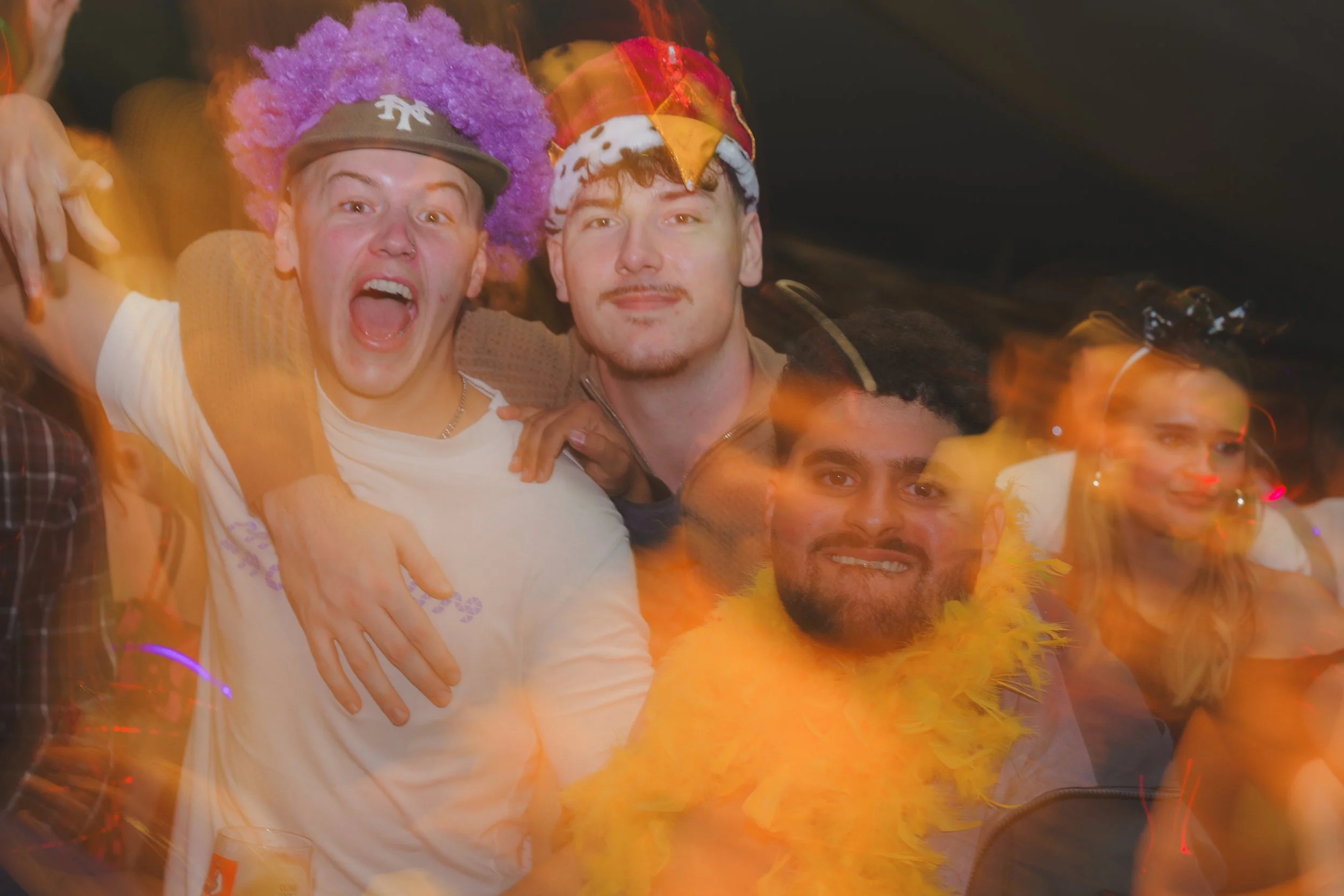 Two men celebrating at a party, one with a purple afro wig and a New York Yankees cap, and the other wearing a colorful jester hat. Both are smiling and appear to be having fun.