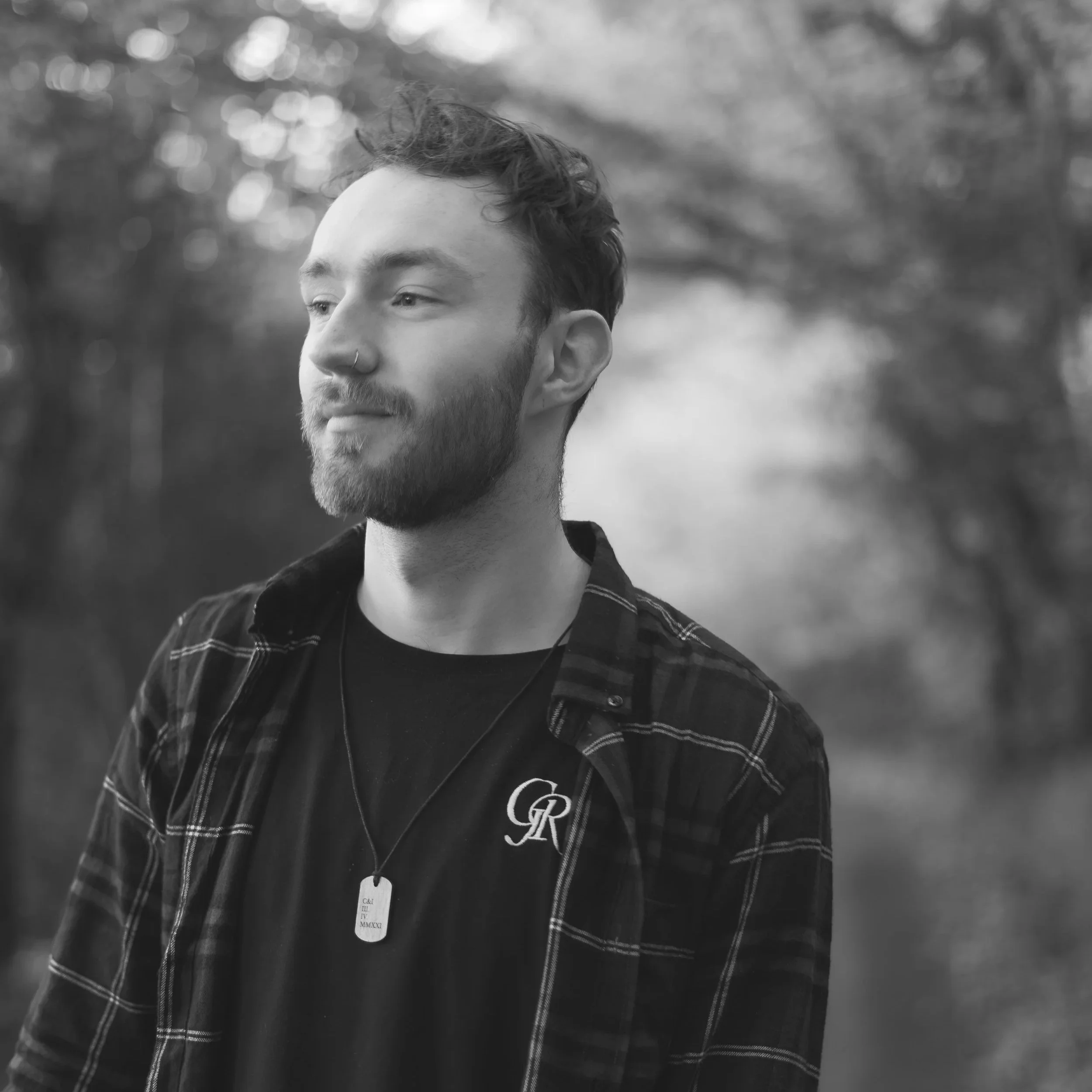 A young man with short hair, a beard, and a nose ring, wearing a plaid shirt over a black shirt with a logo, standing outdoors with blurred trees in the background.