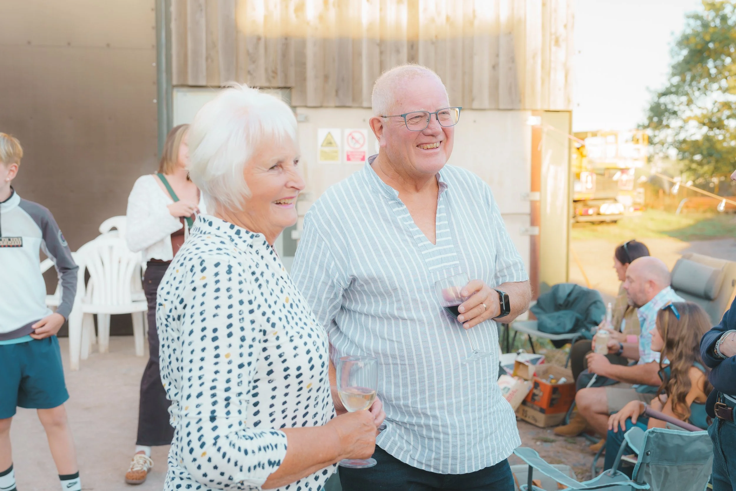 A group of people at an outdoor gathering, smiling and chatting, with two older adults in the foreground holding wine glasses.