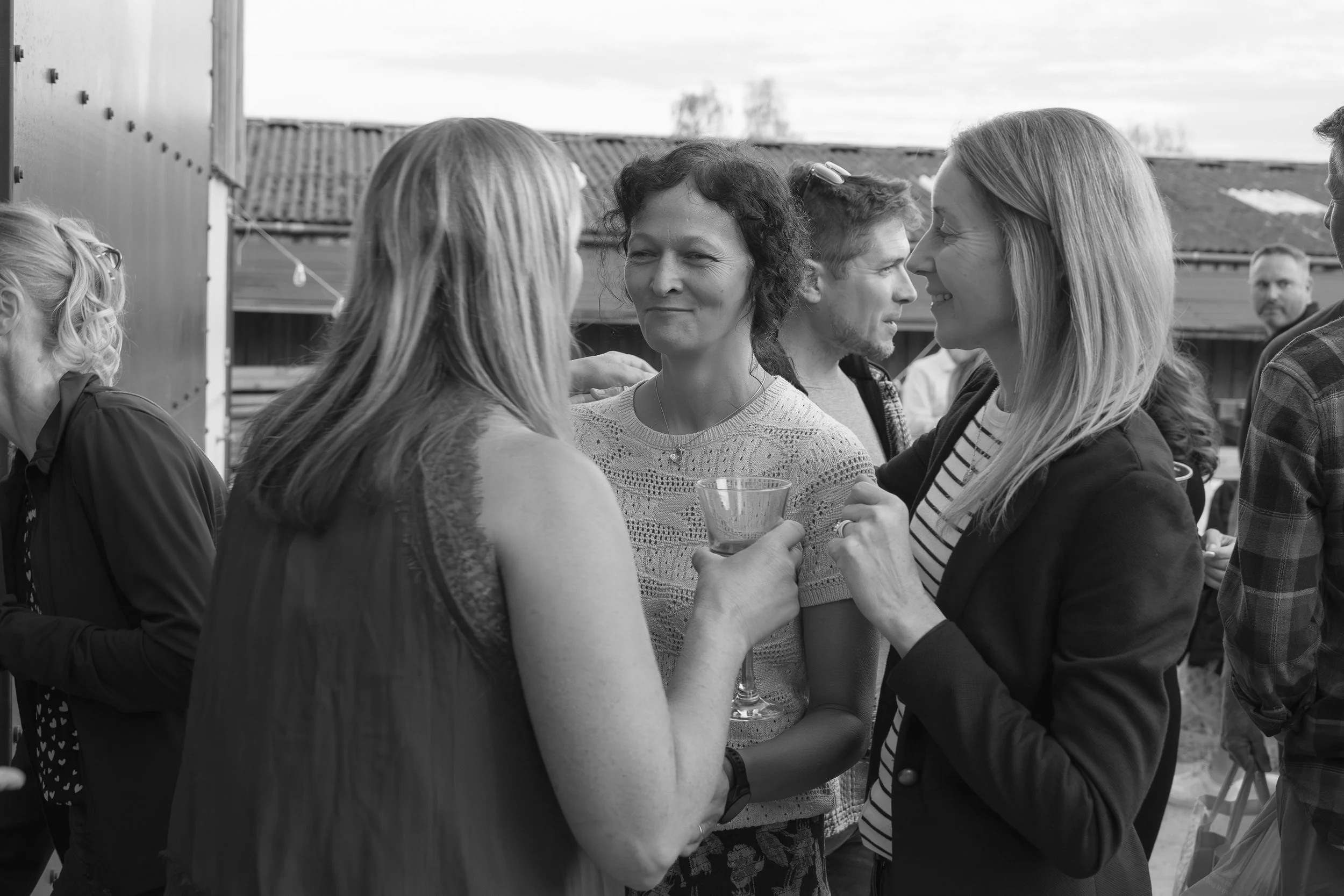 Black and white photo of a group of people conversing at an outdoor gathering, with some holding drinks and smiling.