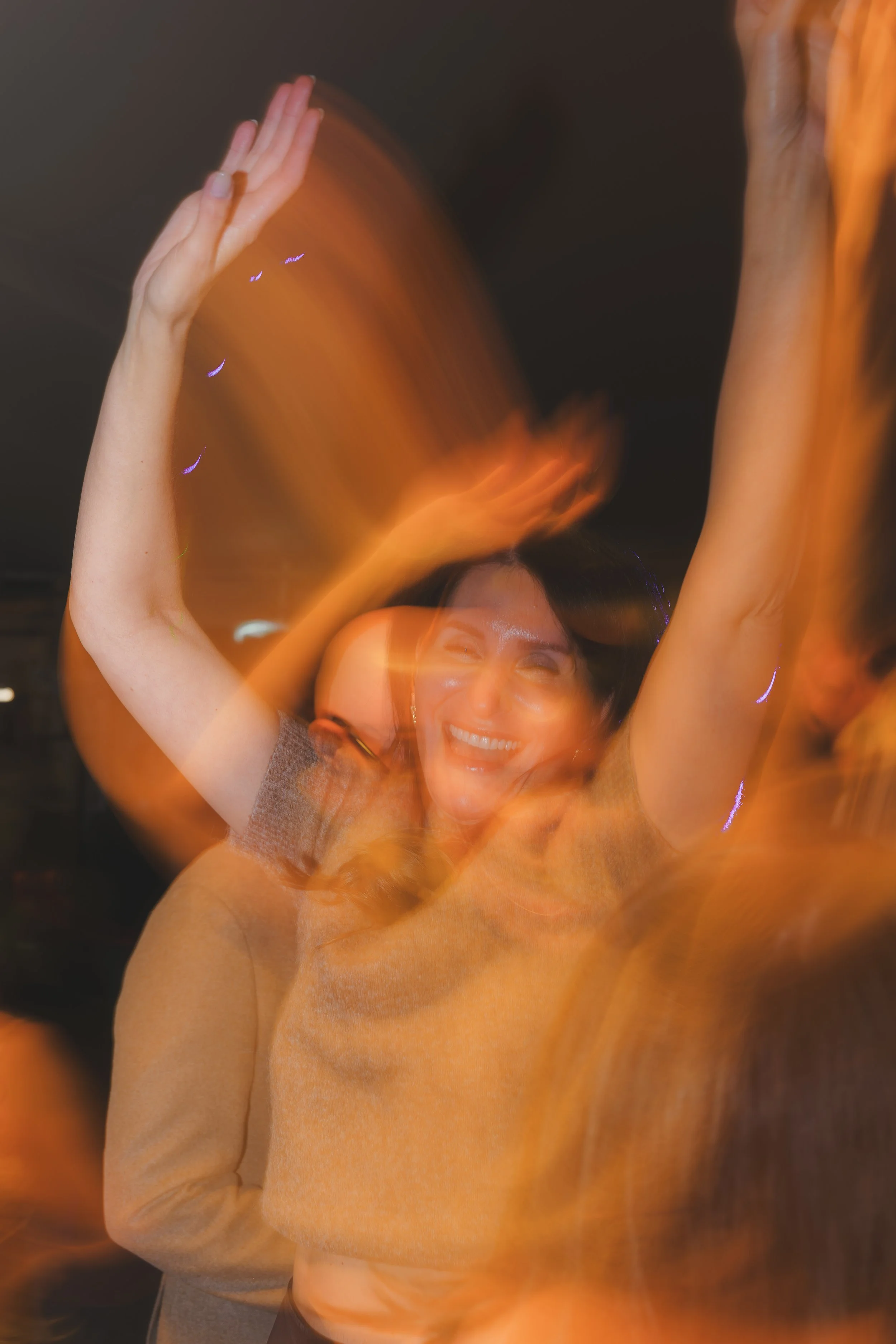A woman smiling and waving with her right hand, her face partially covered by orange light streaks that create a dynamic, blurry effect.