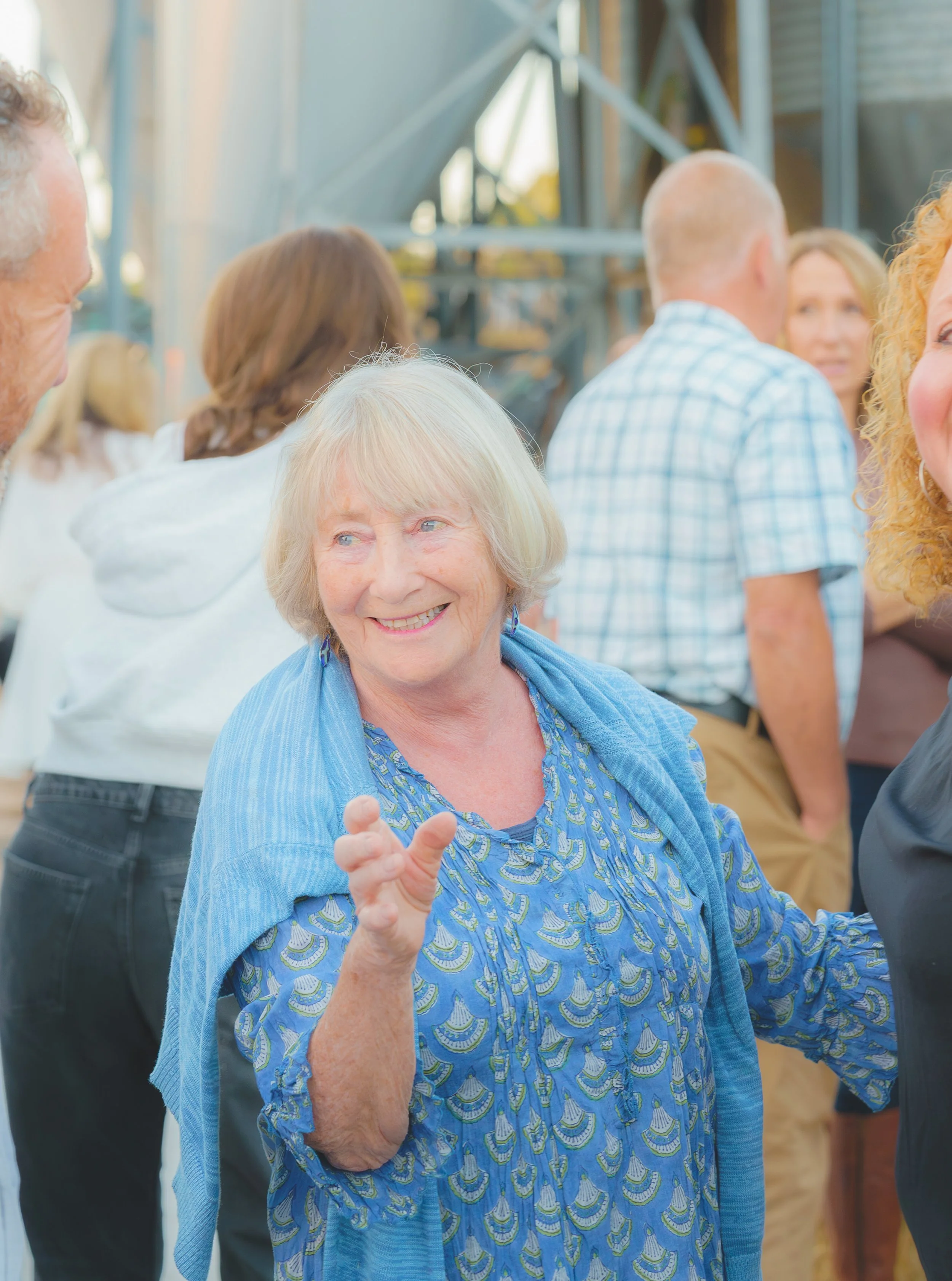 An elderly woman with white hair smiling and gesturing with her hand, wearing a blue patterned blouse and a matching blue shawl, in a social setting with other people in the background.