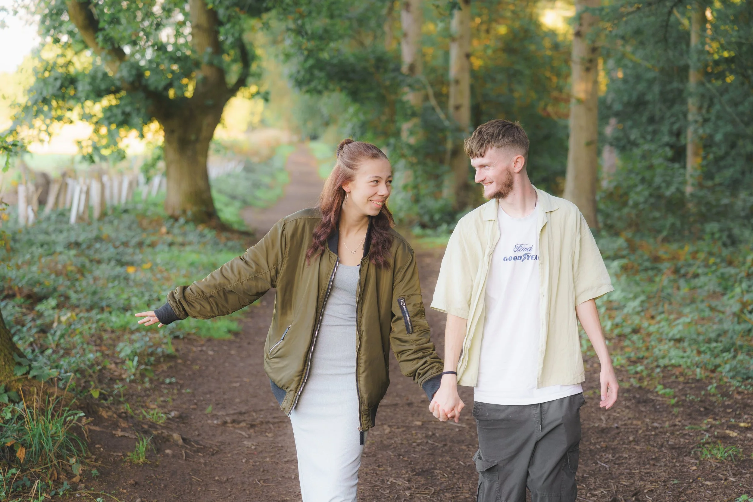A couple holding hands and walking through a forest trail surrounded by trees, with sunlight filtering through the leaves.