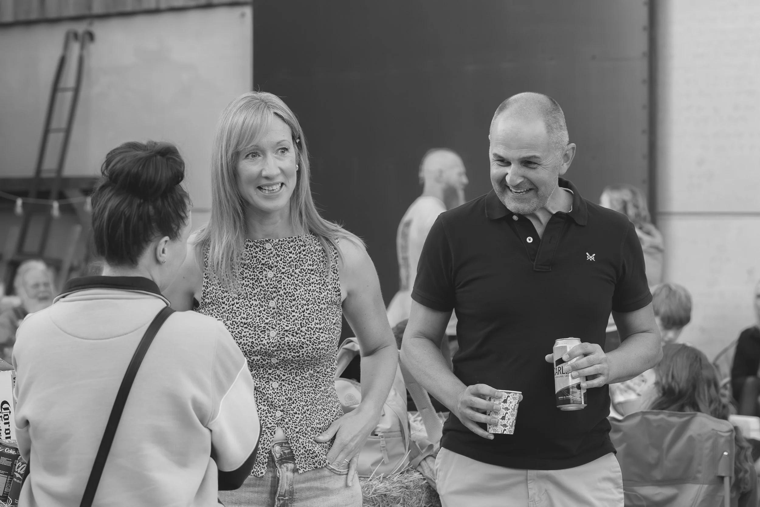A black and white photo shows three people standing and talking at an outdoor event. One woman with long blonde hair, wearing a patterned sleeveless top, is smiling and talking to a woman with dark hair tied in a bun, wearing a light-colored top. A m