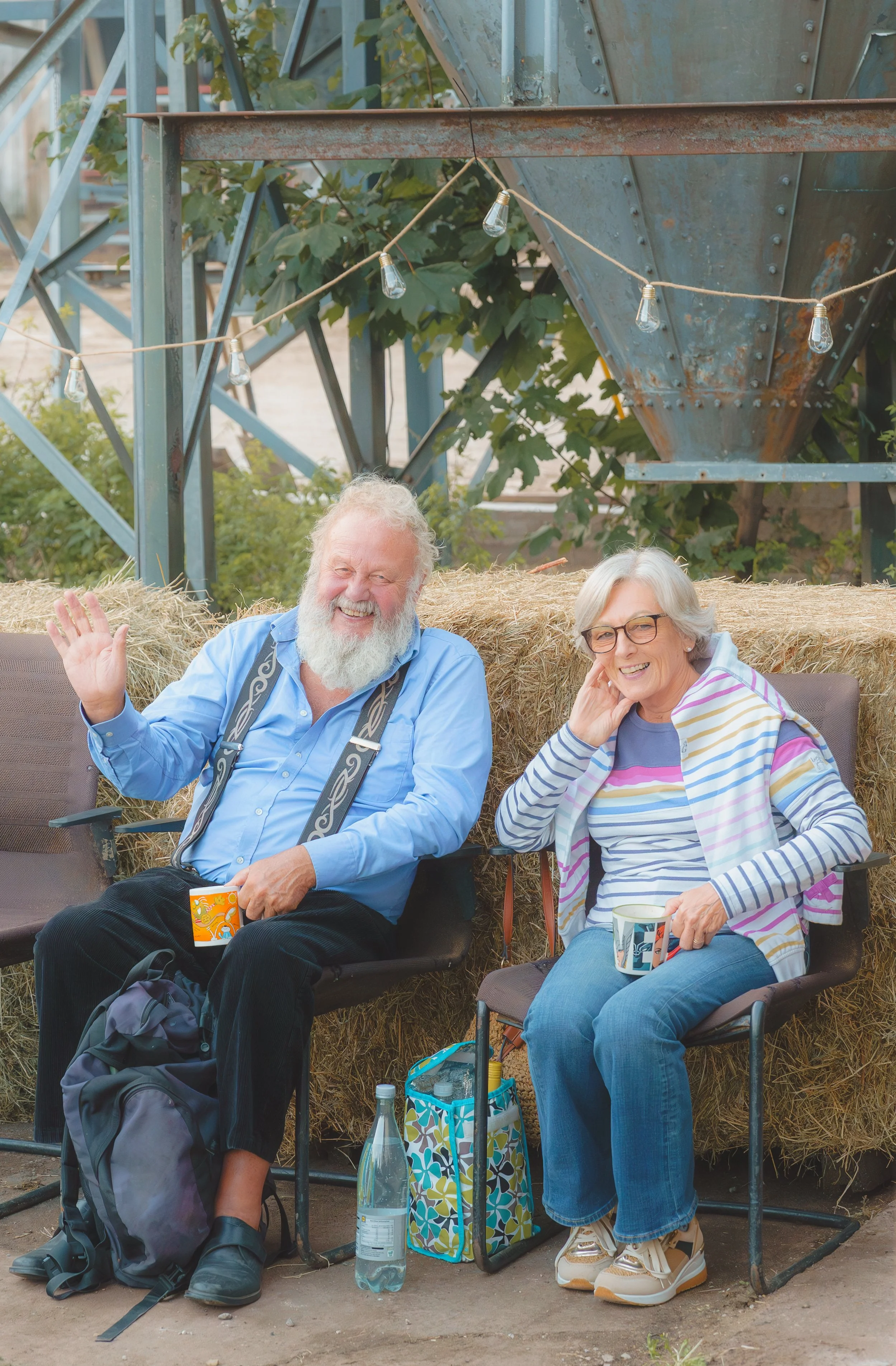 Smiling elderly man in a blue shirt and woman in a striped shirt sitting on chairs outdoors, with hay bales behind them and string lights hanging above, in a rustic setting.
