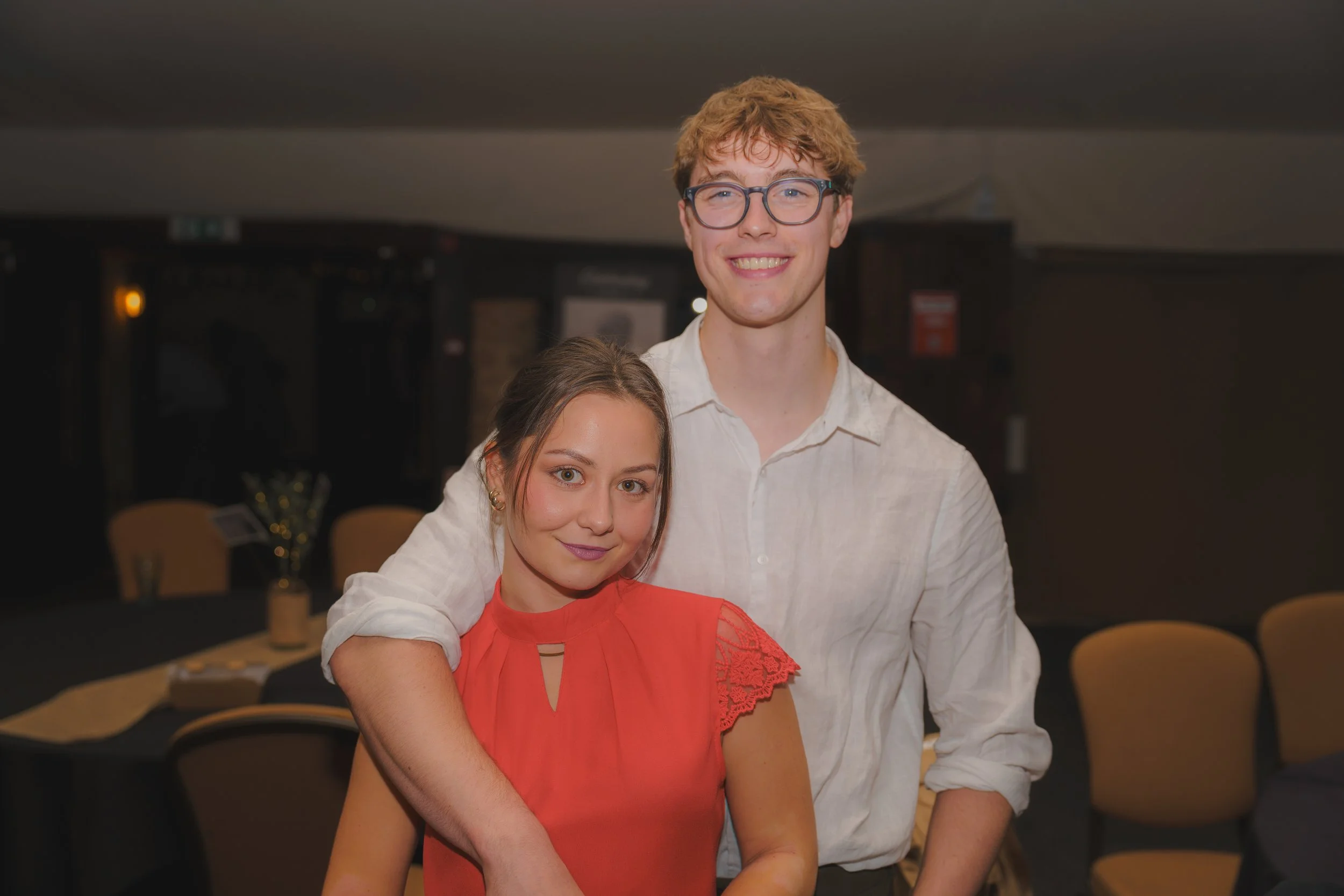 A young woman in a red dress and a young man with glasses and a light shirt stand close together in a dimly lit restaurant or event space, smiling at the camera.