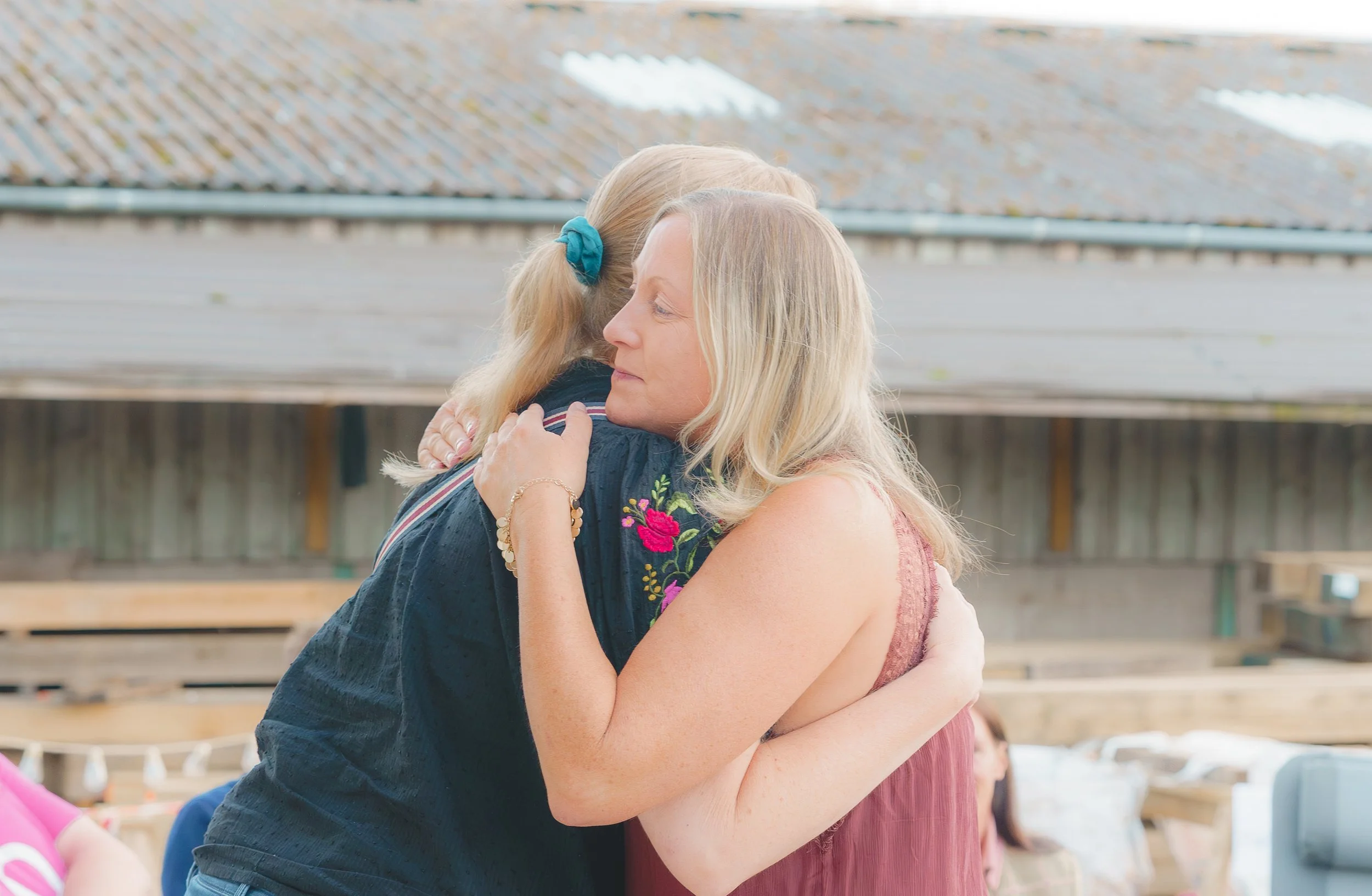 Two women hugging each other at an outdoor gathering. One woman has blonde hair, and the other is partially turned away with a ponytail.