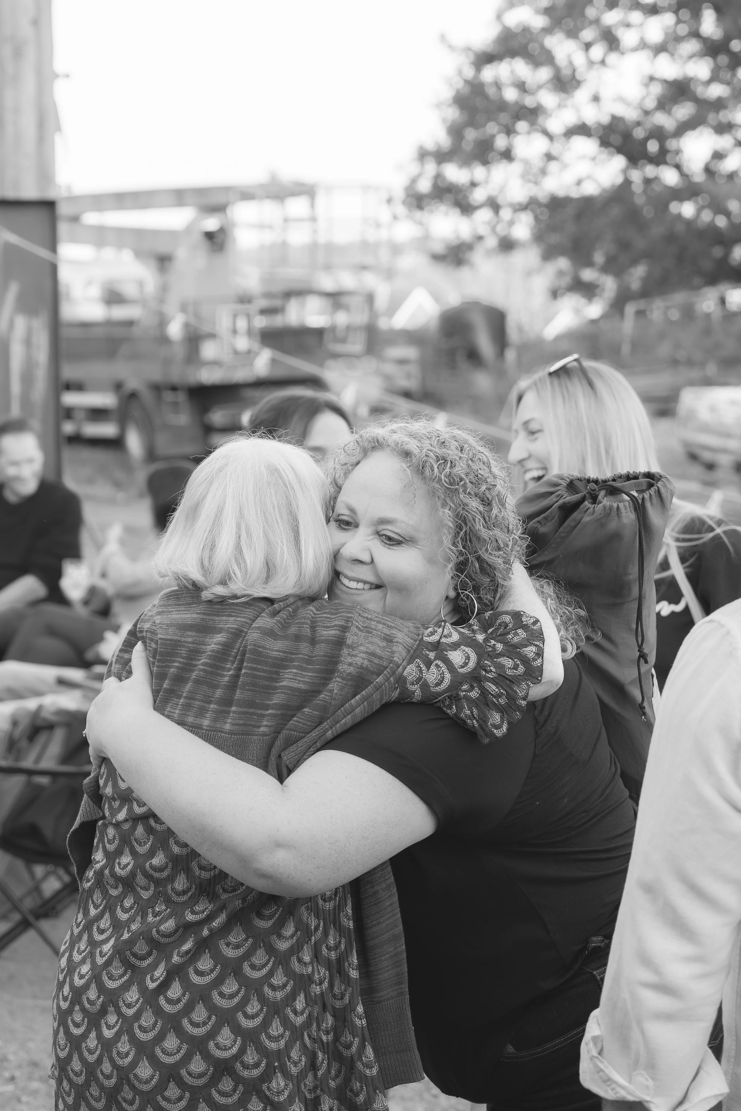 A group of women hugging and smiling outdoors, with other people and trees in the background