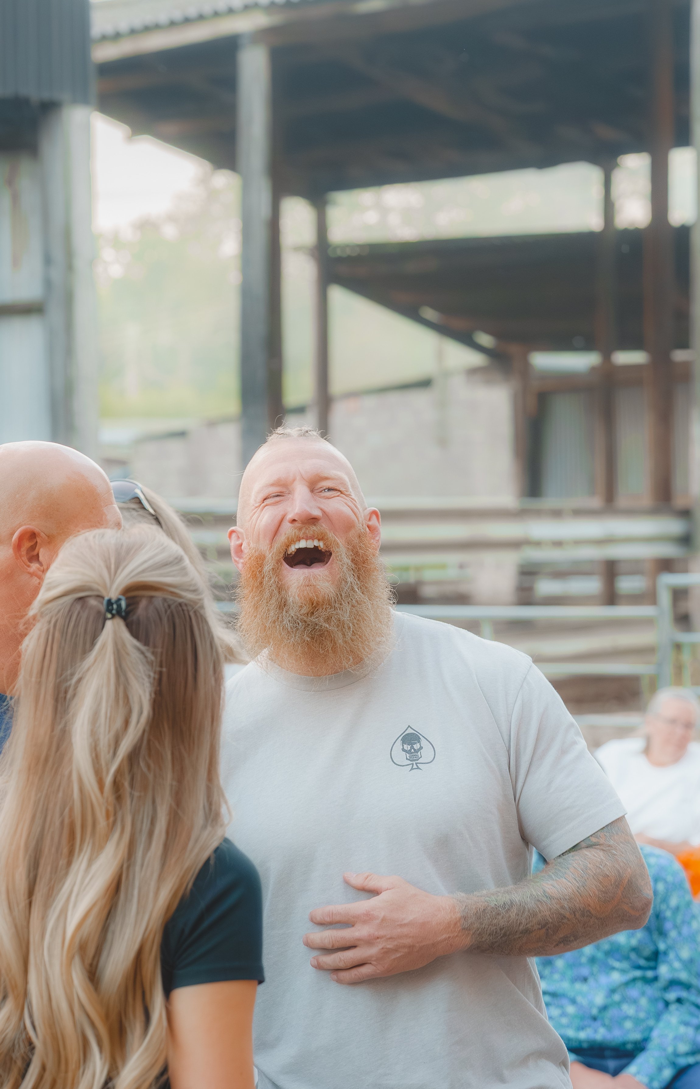 A group of people socializing outdoors, with one bearded man wearing a white T-shirt laughing and smiling, surrounded by others including a woman with long blonde hair and another bald man.