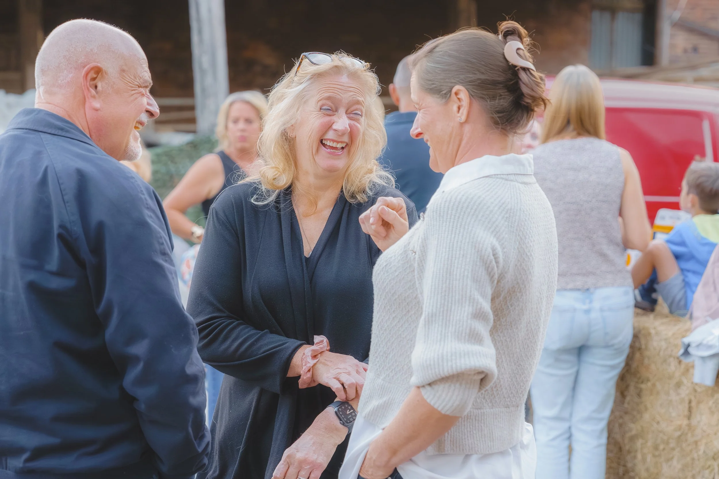 Three adults laughing and holding hands during a gathering outdoors, with a group of people in the background.