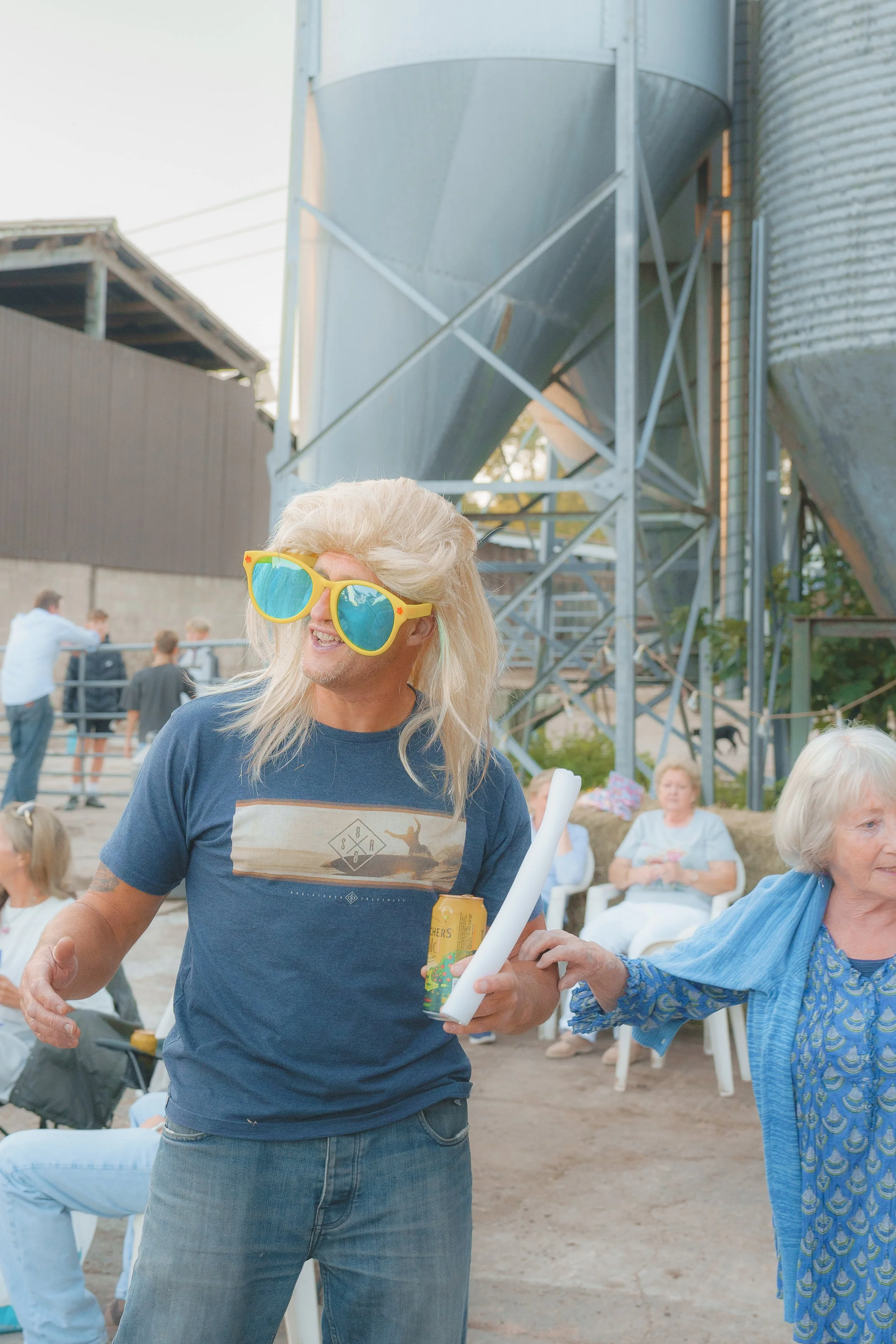 A man with long blonde hair wearing bright yellow sunglasses and a blue t-shirt talking to an older woman in a blue dress during an outdoor gathering.