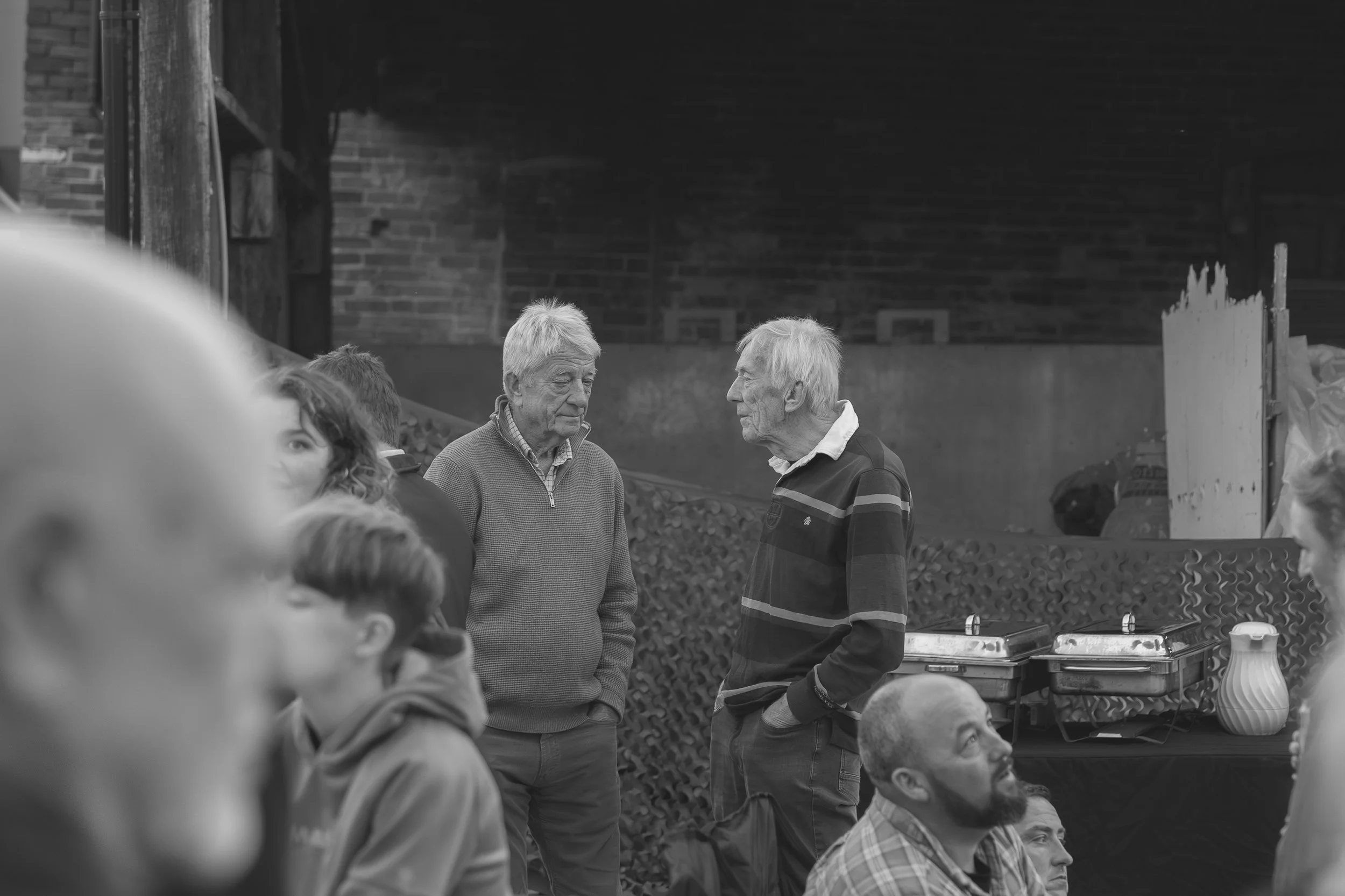 Two elderly men engaged in conversation at a social gathering outdoors, with several other people partially visible and a brick building in the background.