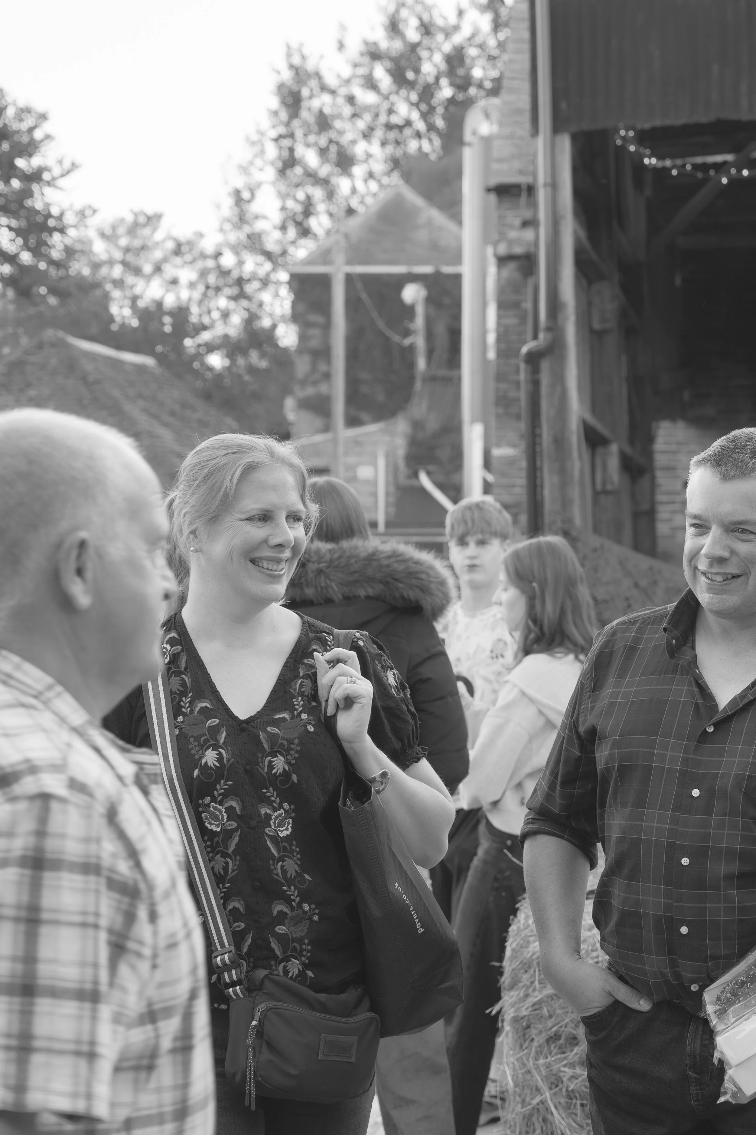 People gathered together outdoors, engaging in conversation, with a woman smiling and a man in a plaid shirt. There are other individuals in the background, and a rustic building with a sloped roof and string lights, suggesting a casual social event 
