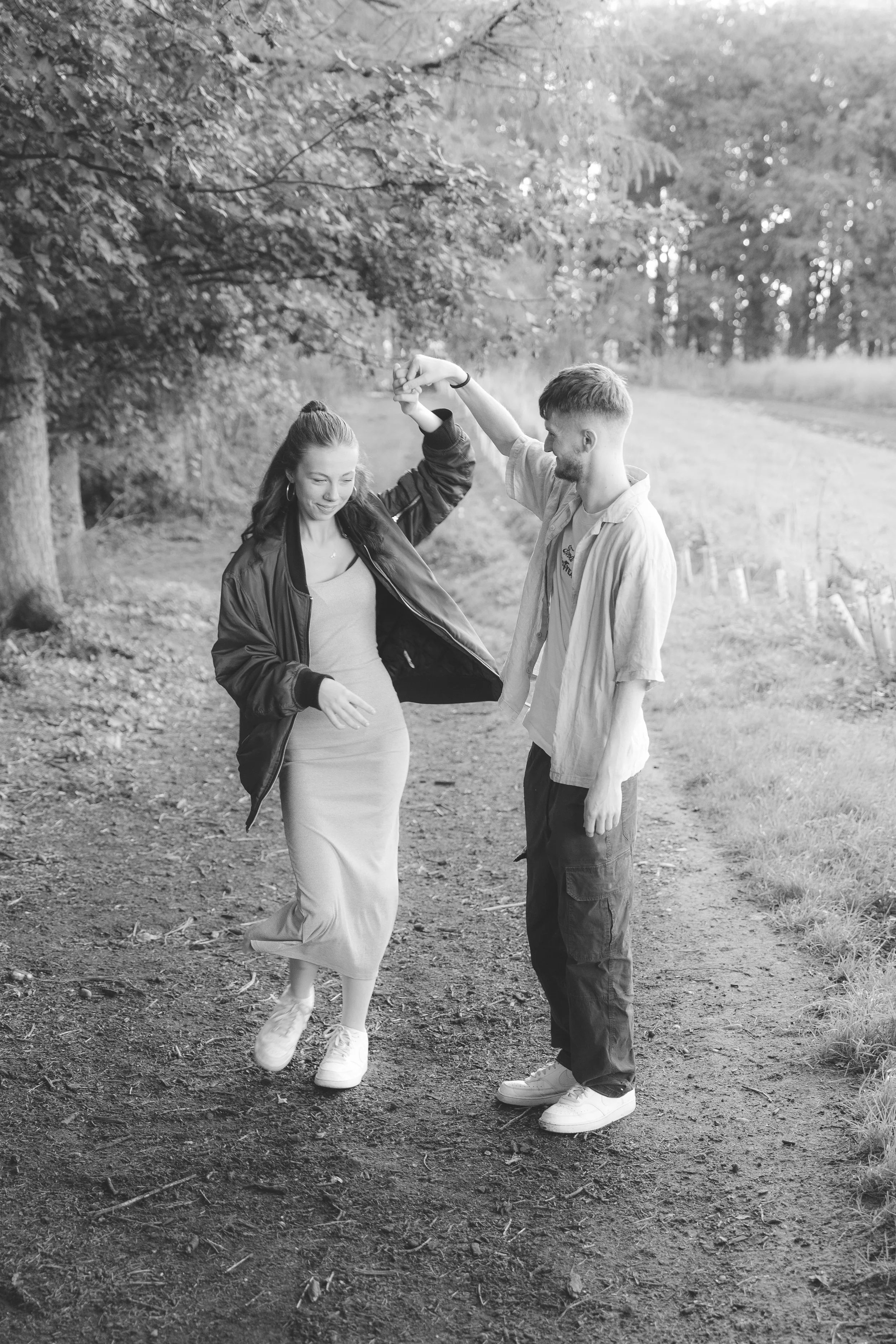 A young woman and a young man are dancing together outdoors on a dirt path in a wooded area. The woman is wearing a dress and a jacket, and the man is wearing a shirt and pants. They are holding hands and smiling, enjoying a moment together.