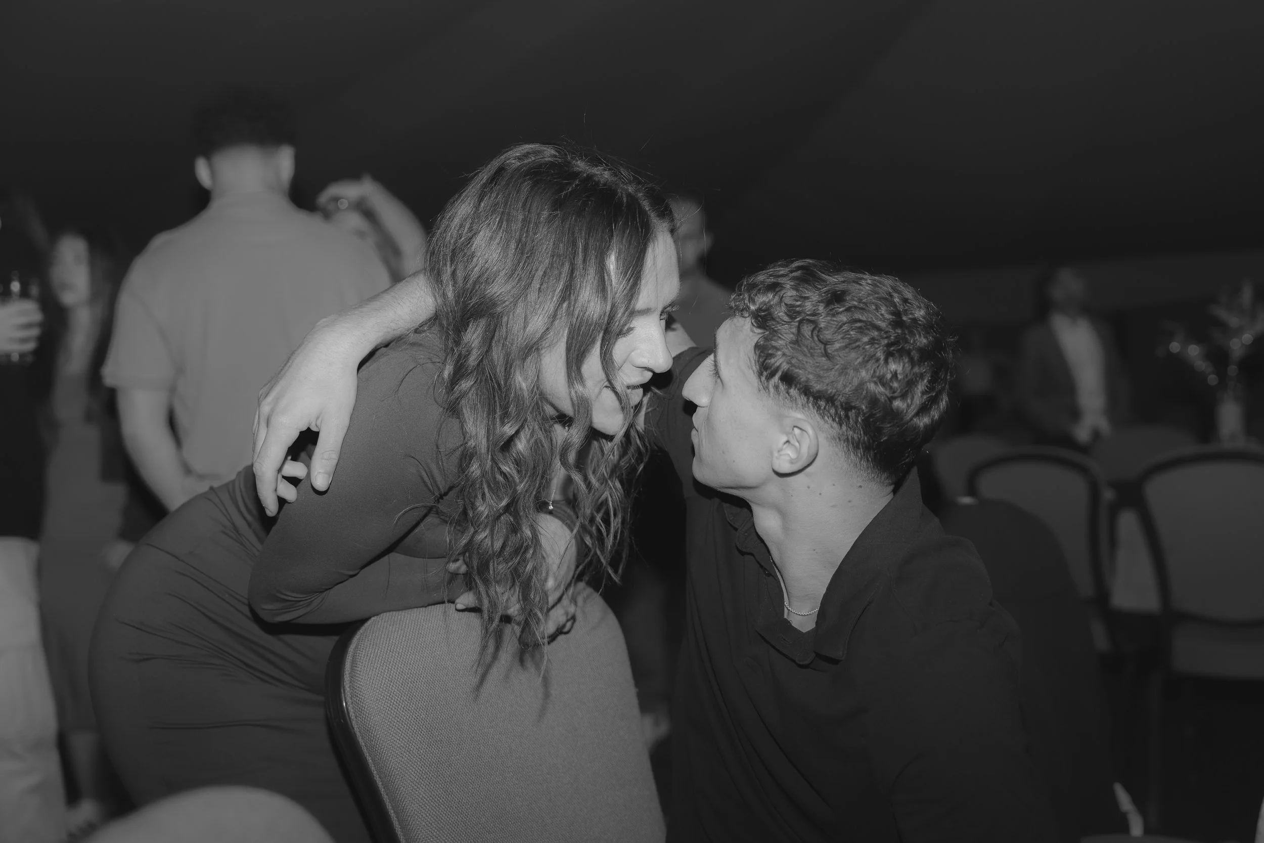 A black-and-white photo of a romantic moment between a young man and woman in a social gathering. The woman has long wavy hair and is leaning close to the young man who has short hair. They are gazing into each other's eyes with their foreheads nearl