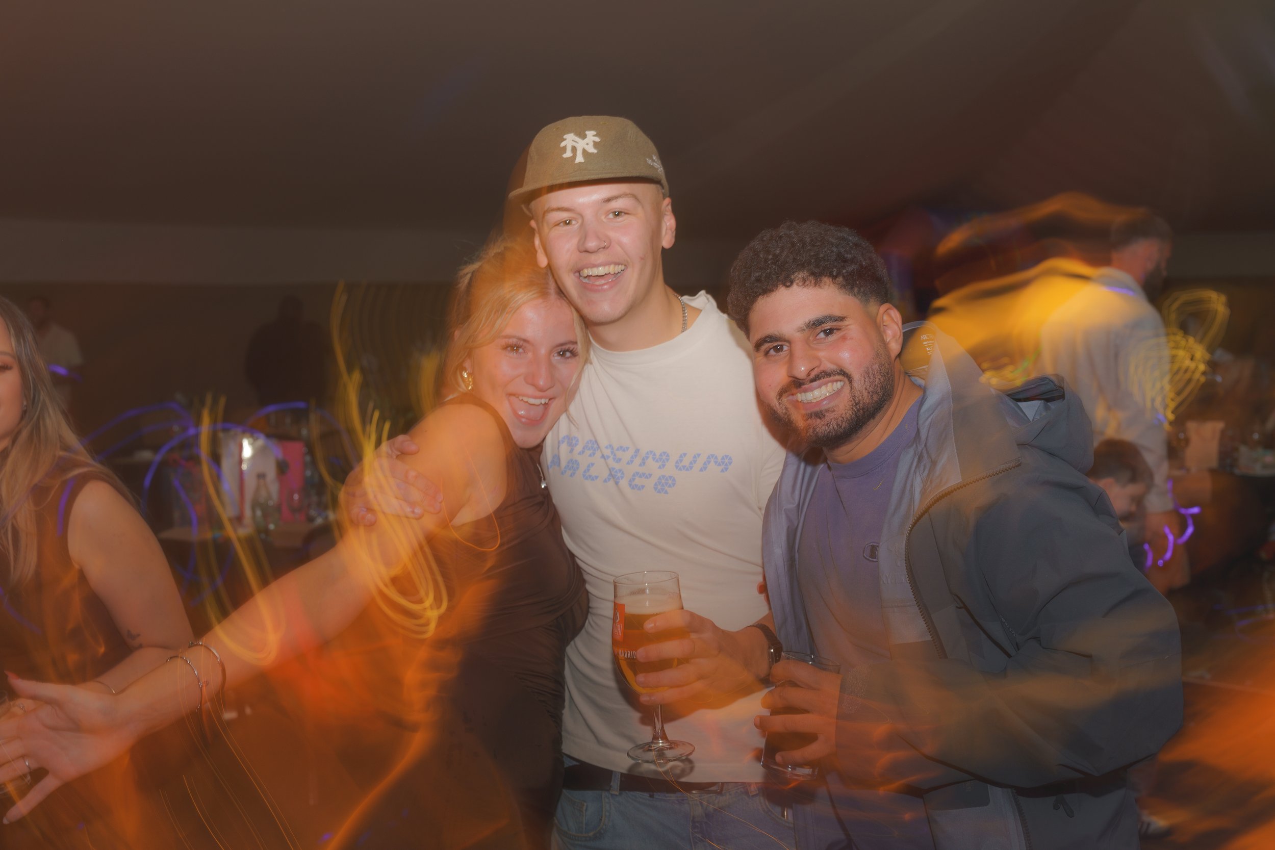 Group of three young adults smiling and posing together at a party, with drinks in hand, in a dimly lit venue.