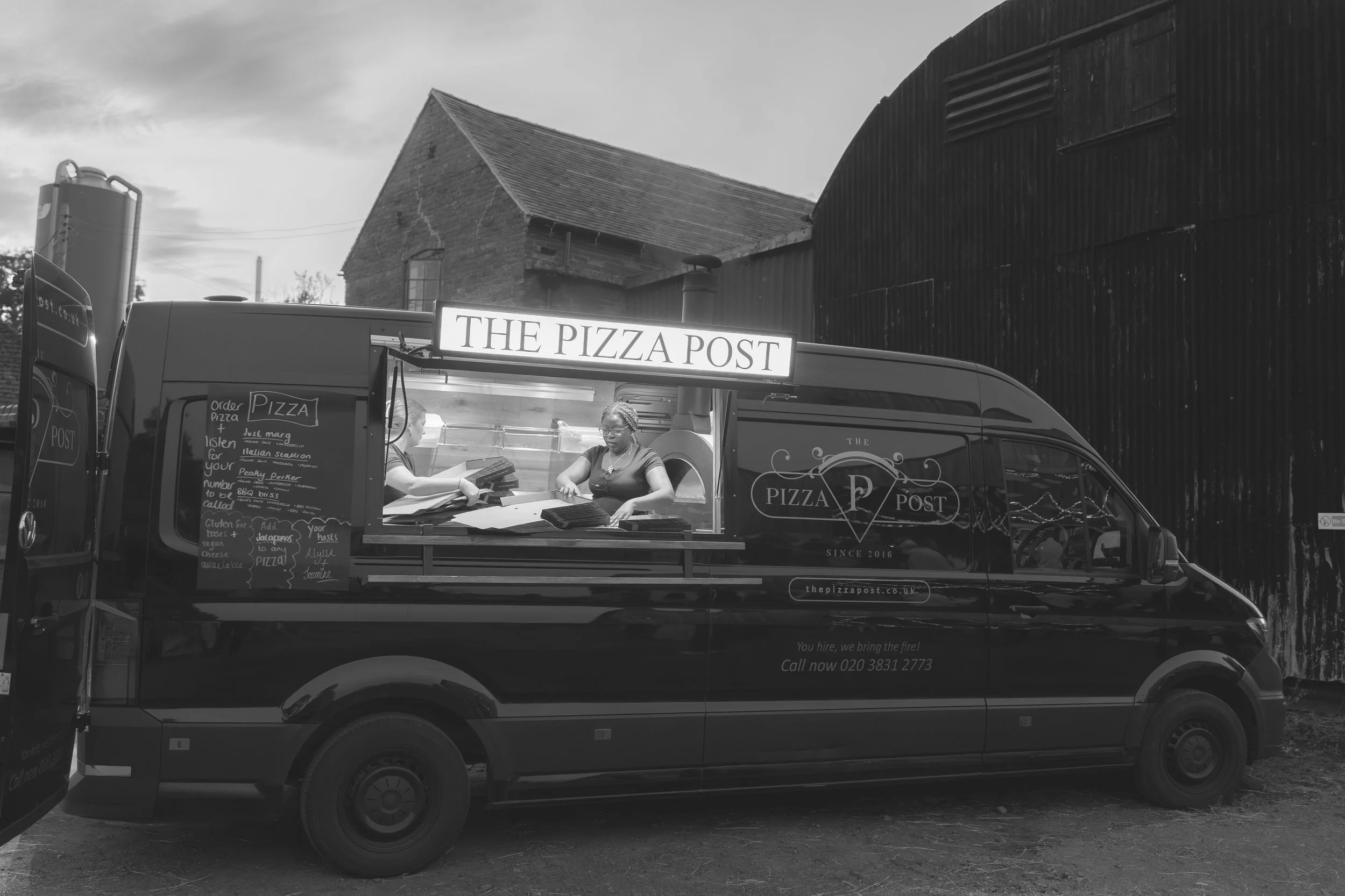 A food truck named 'The Pizza Post' serving pizza, with two women working at the window. The truck is parked outdoors with rustic buildings in the background.