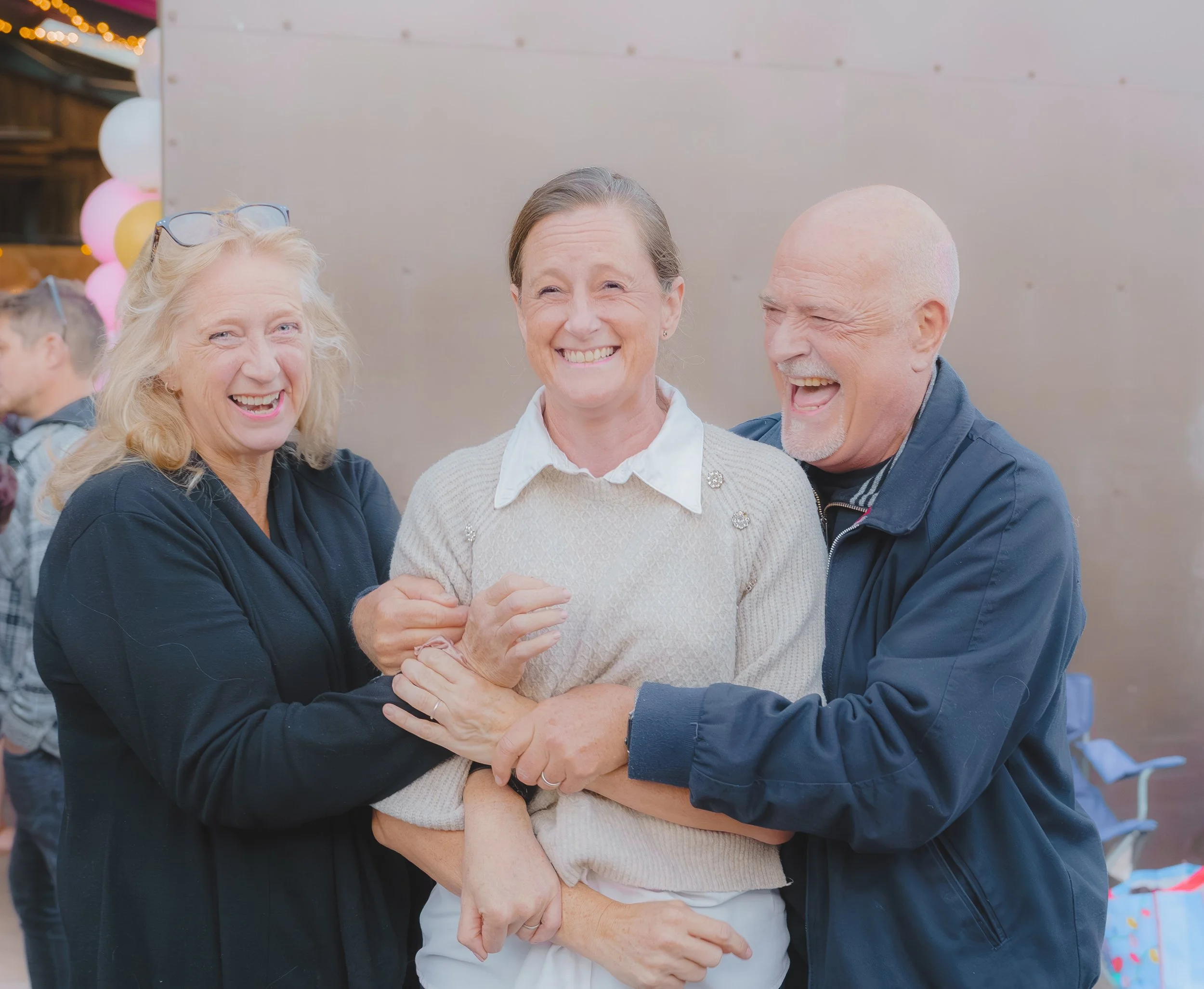 Three people, an elderly woman with glasses on her head, a smiling middle-aged woman, and an elderly man, are hugging and laughing at an outdoor gathering with balloons in the background.