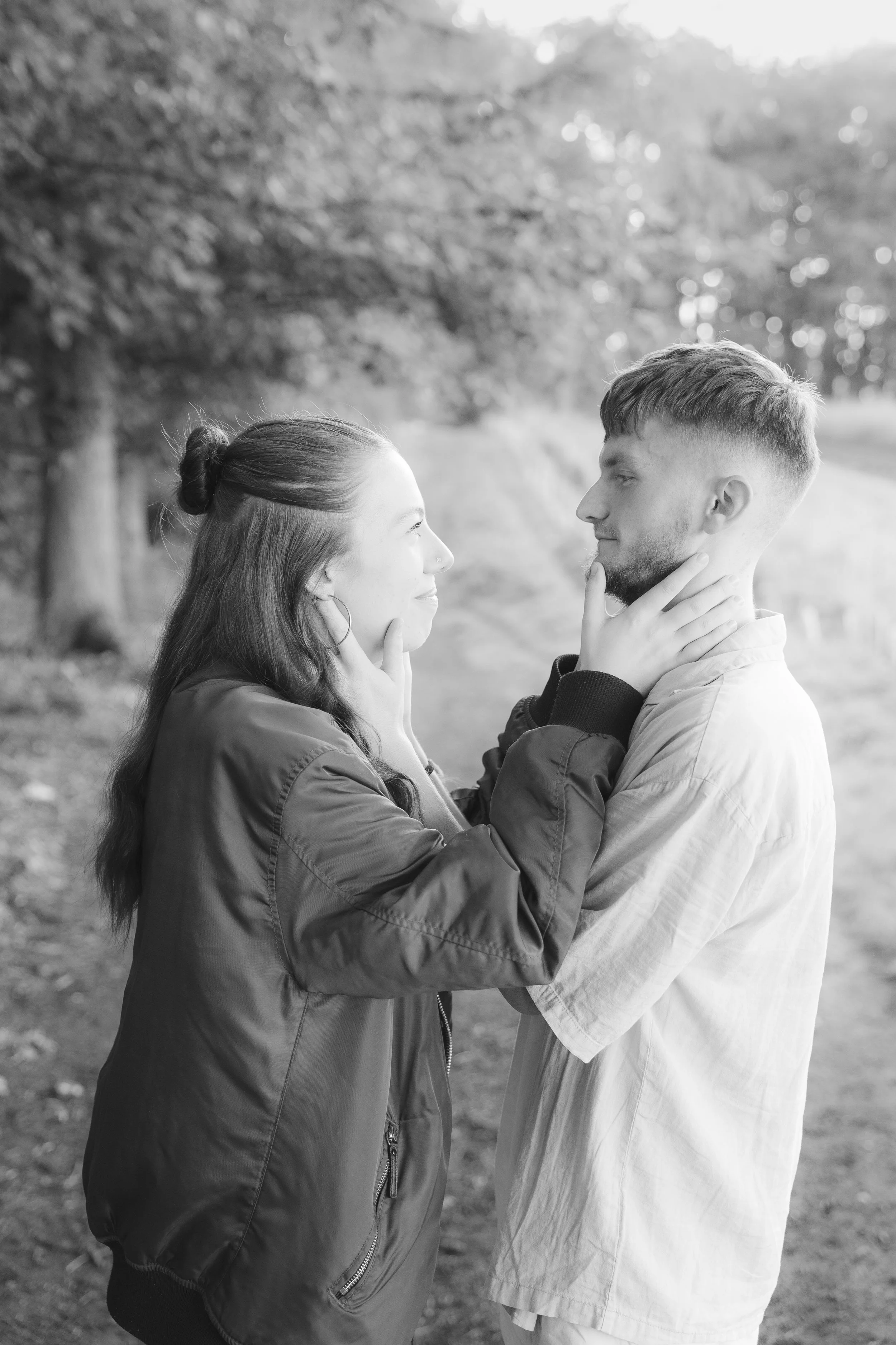 A black-and-white photo of a young couple standing outdoors, gazing into each other's eyes, with the woman touching the man's face and the man holding her waist, surrounded by trees and foliage.