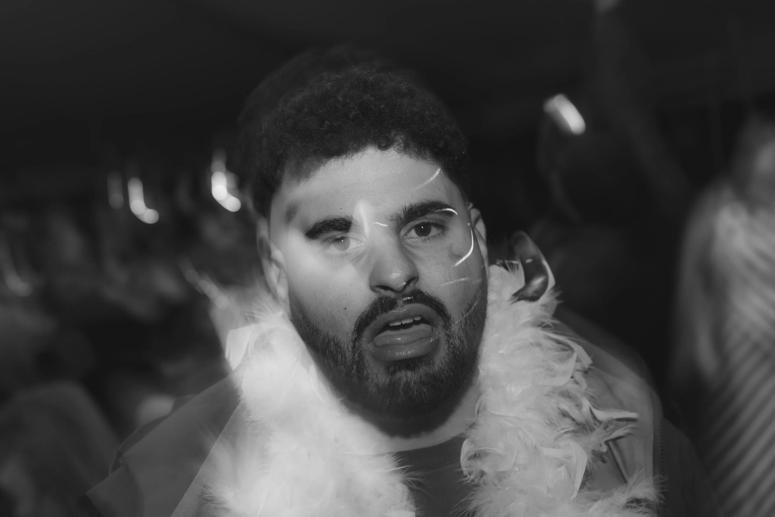 Black and white close-up of a young man with curly hair and a beard, wearing a feathered or furry outfit, looking directly at the camera with one eye winking.