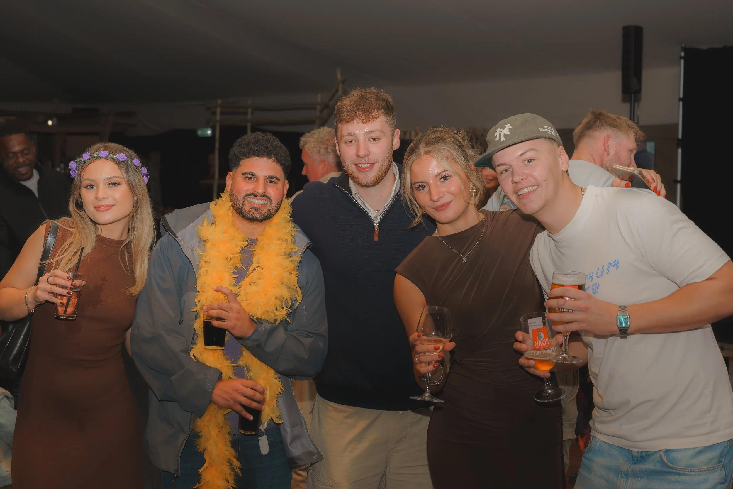 Group of five young adults at a celebration, smiling and holding drinks, with some wearing festive accessories like a purple floral headband and a yellow feathered boa.