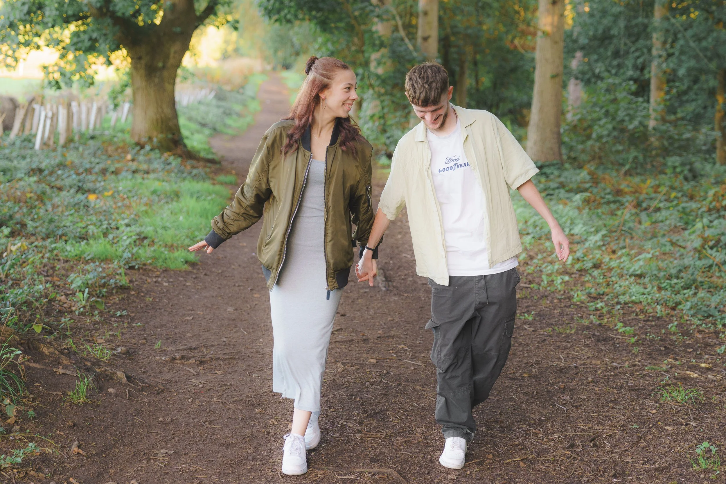 A young woman and a young man walking hand in hand on a dirt path through a wooded area with green trees and foliage, smiling and enjoying each other's company.