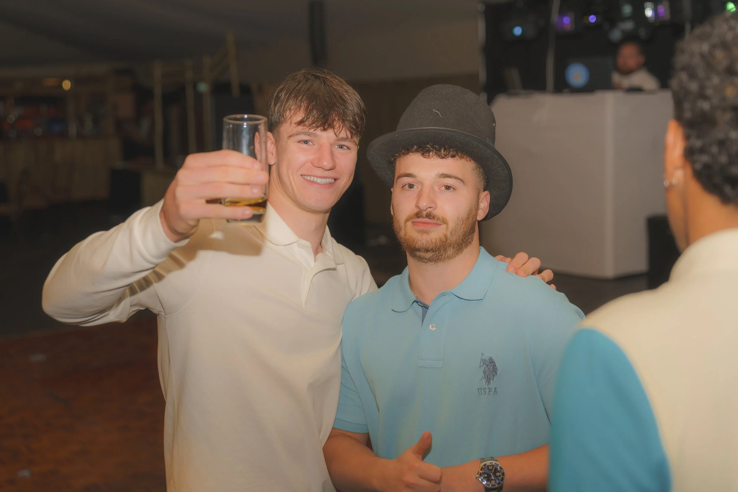 Two men smiling and posing for a photo at a celebration, one holding a glass of drink. One man is wearing a white shirt, and the other is wearing a light blue polo shirt and a black top hat. There are other people and a DJ setup in the background.