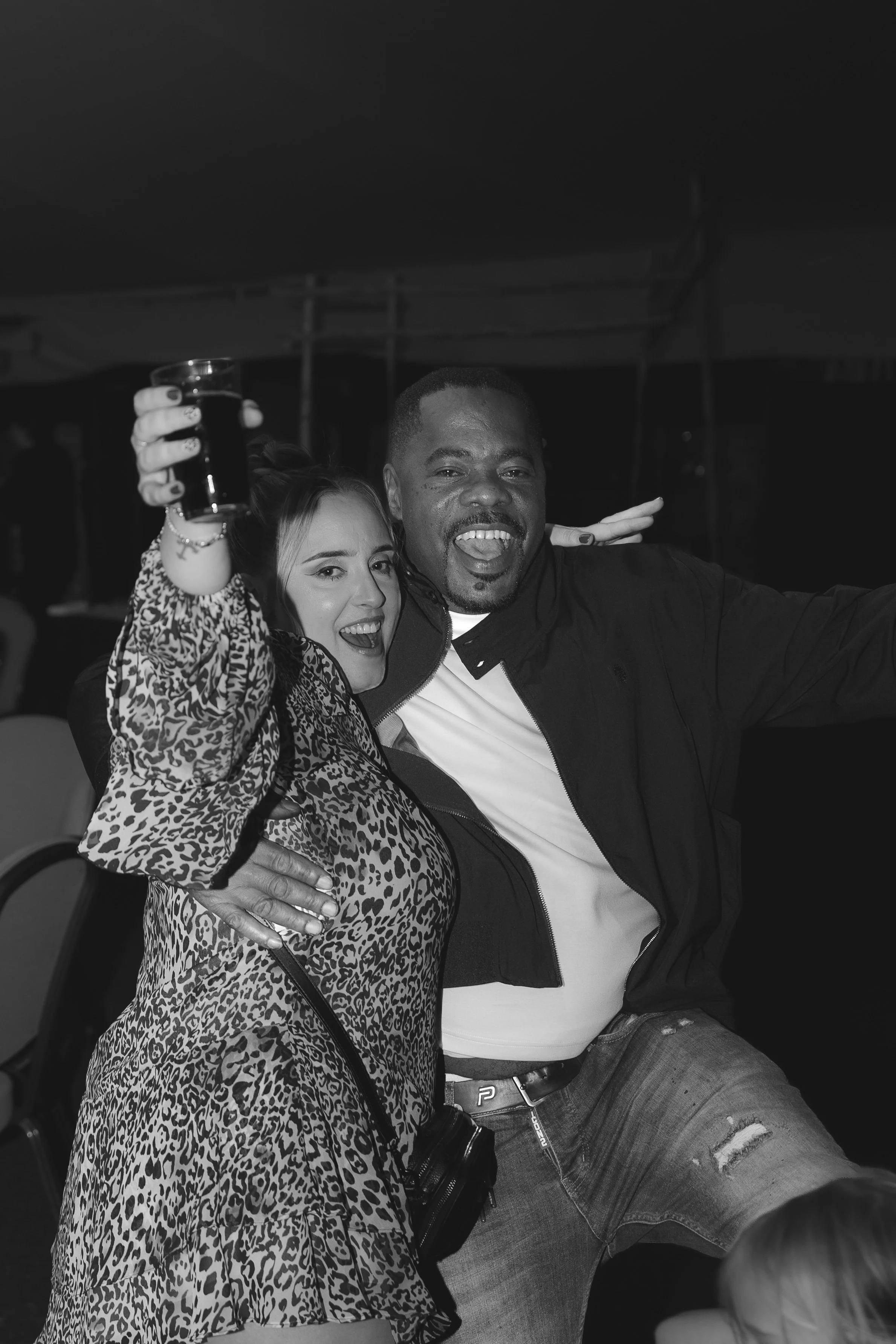 A black and white photo of a woman and man celebrating and smiling at a party. The woman, wearing a leopard print dress, holds a drink in her hand. The man, wearing a white t-shirt and black jacket, has his tongue out and is making a peace sign with his fingers. They are close together, looking happy, with a dark and out-of-focus background.