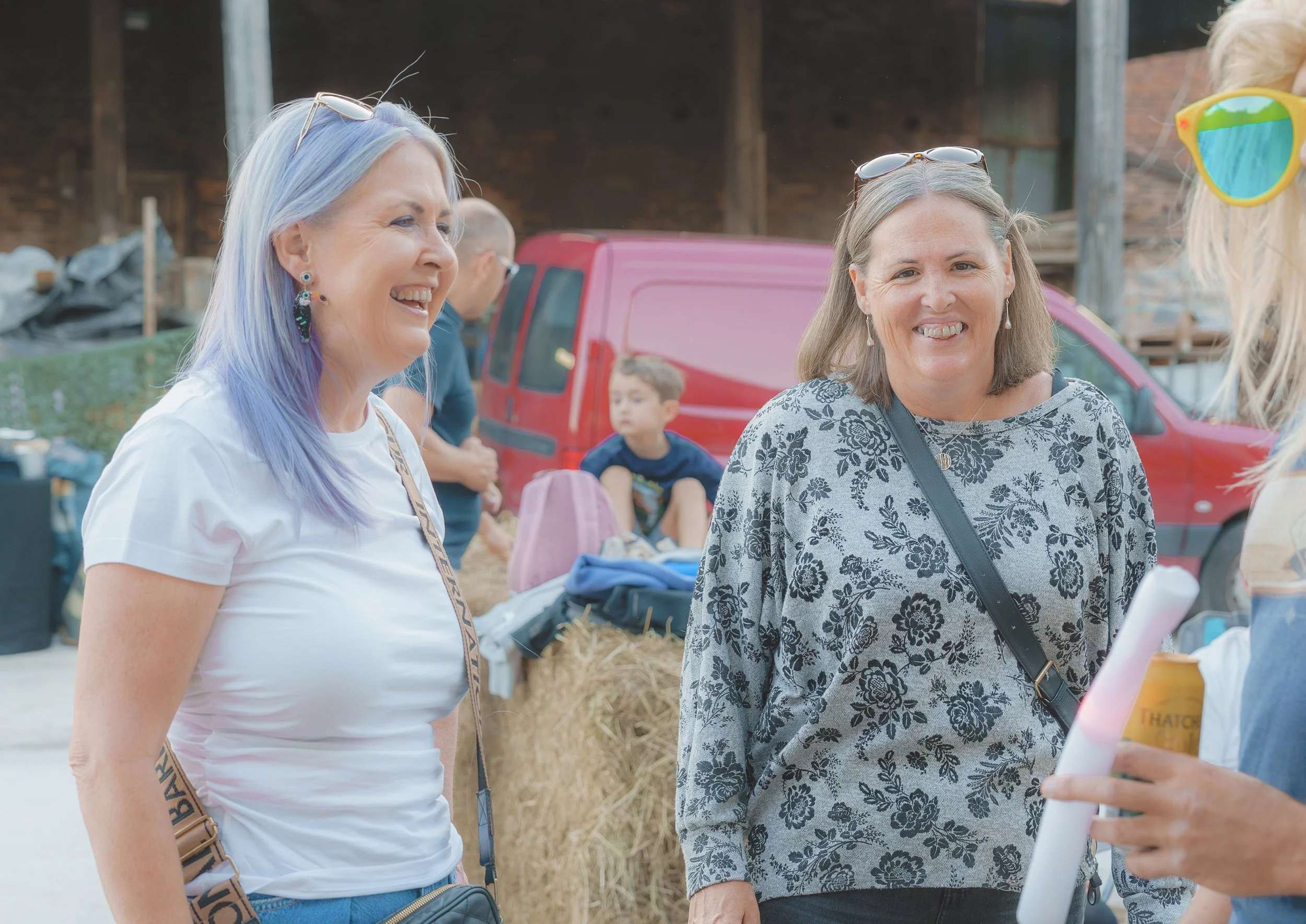 Two women smiling and talking outdoors at a fair or market, with a young boy in the background, a red vehicle, and a wooden structure.