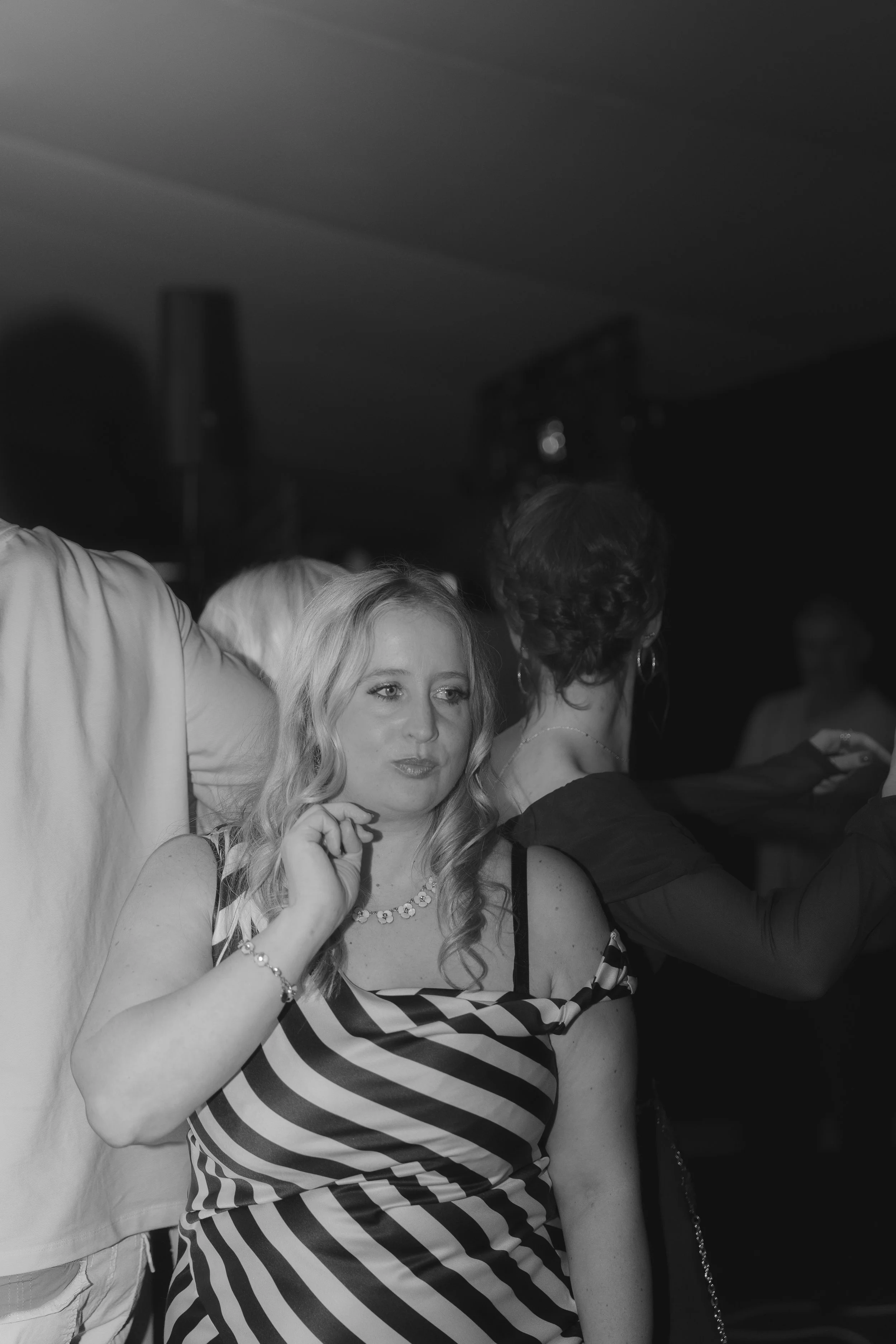 A woman with long, wavy hair and a striped dress, wearing a necklace and bracelet, smiling and holding her hand near her face at a party or social event, with other people visible in the background.