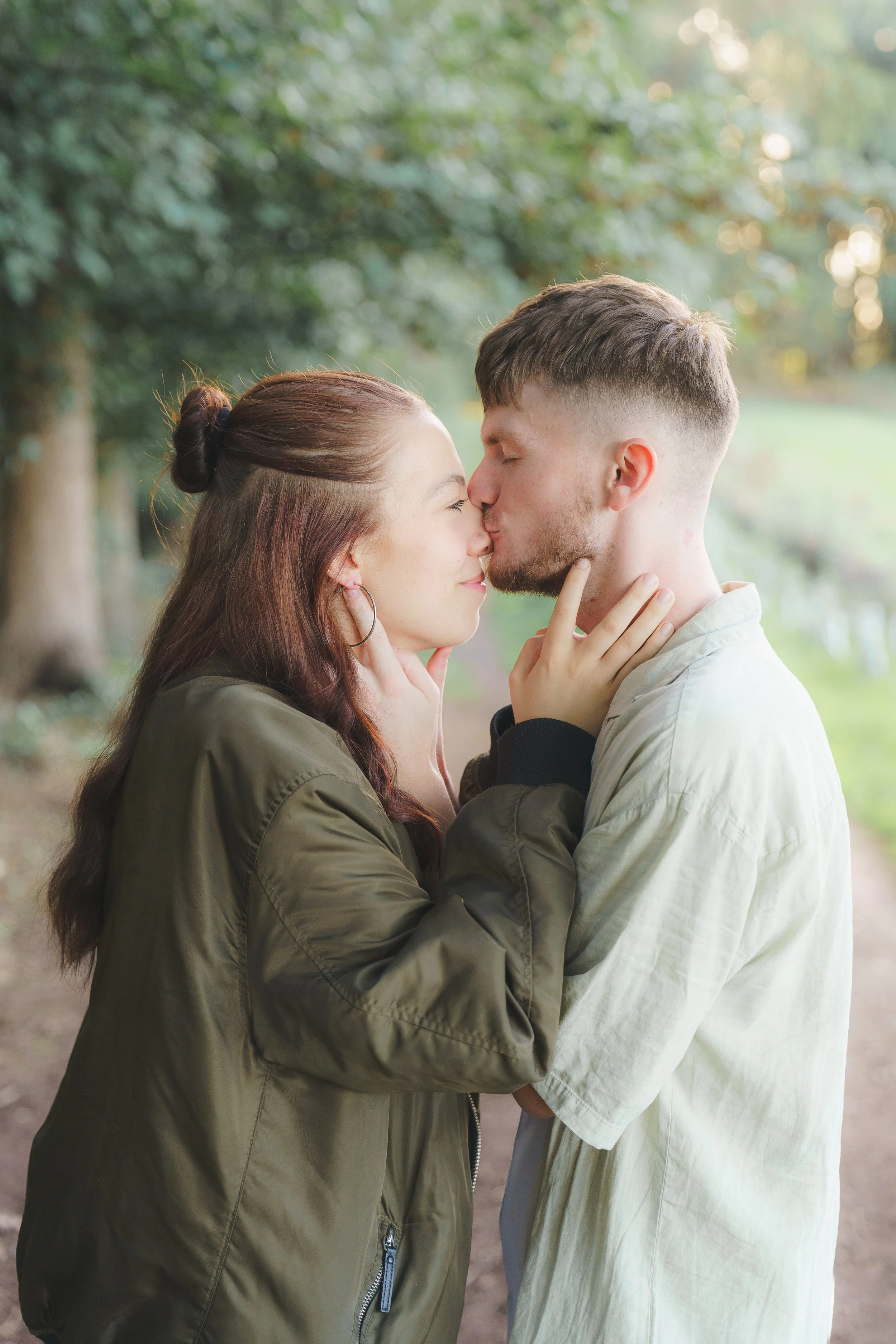 A young woman and man are kissing outdoors in a wooded area, their hands gently touching each other's faces, with trees and sunlight in the background.