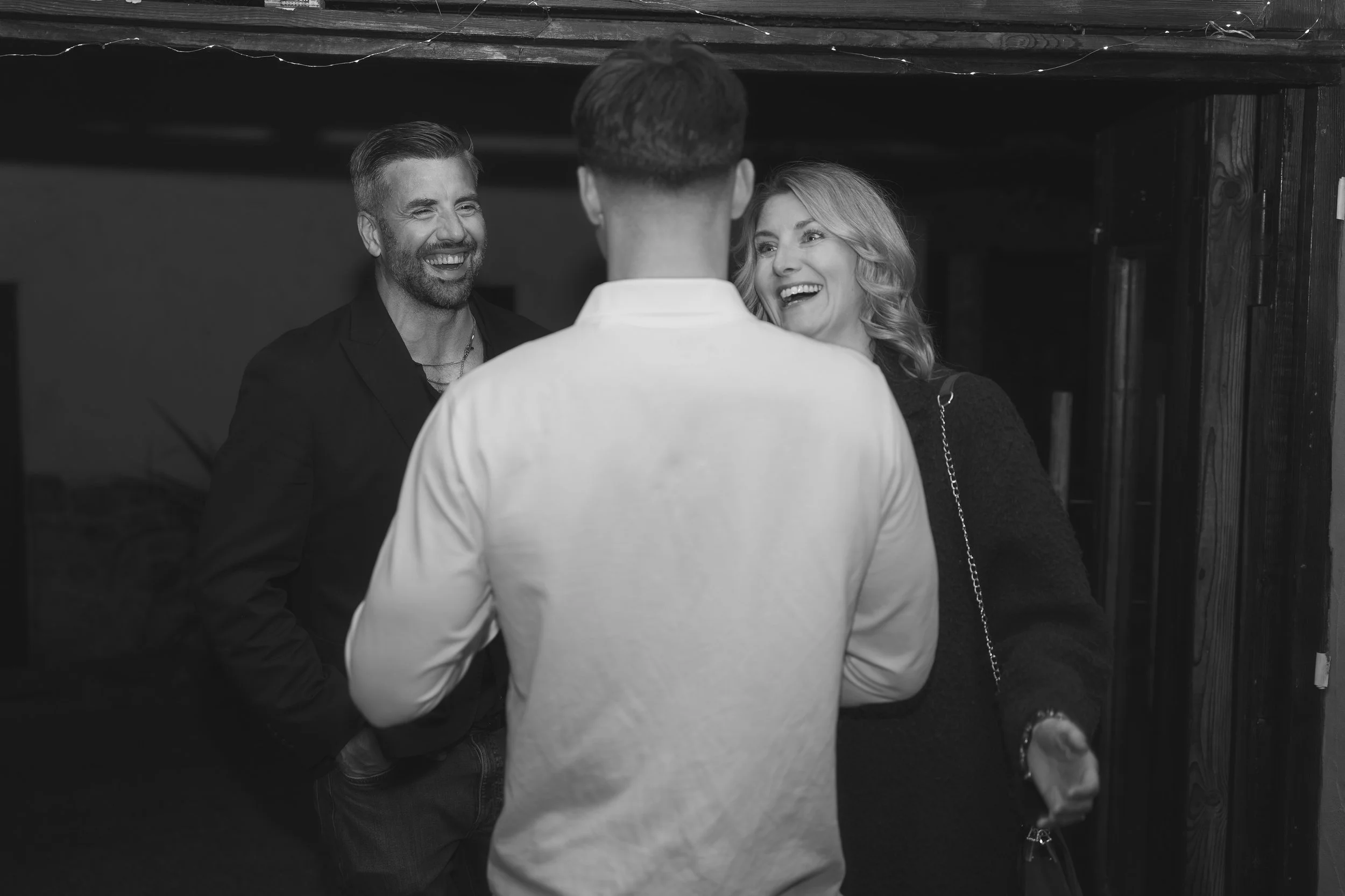 Three people are standing close together, smiling and engaged in conversation at an indoor social gathering. The person facing away from the camera is wearing a light-colored shirt, while the other two are facing the camera, with one man in a dark bl
