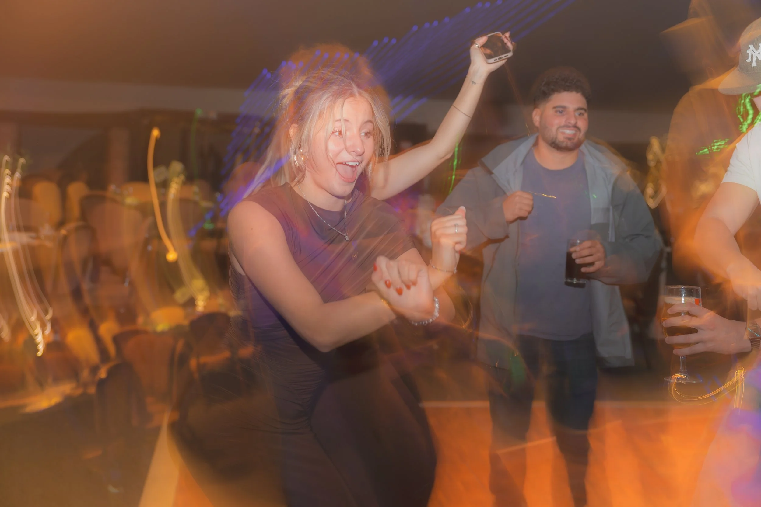 A group of people dancing at a party or celebration, with a blonde woman in the foreground holding a phone high in the air and smiling, and a man in the background holding a drink, all surrounded by colorful lights.