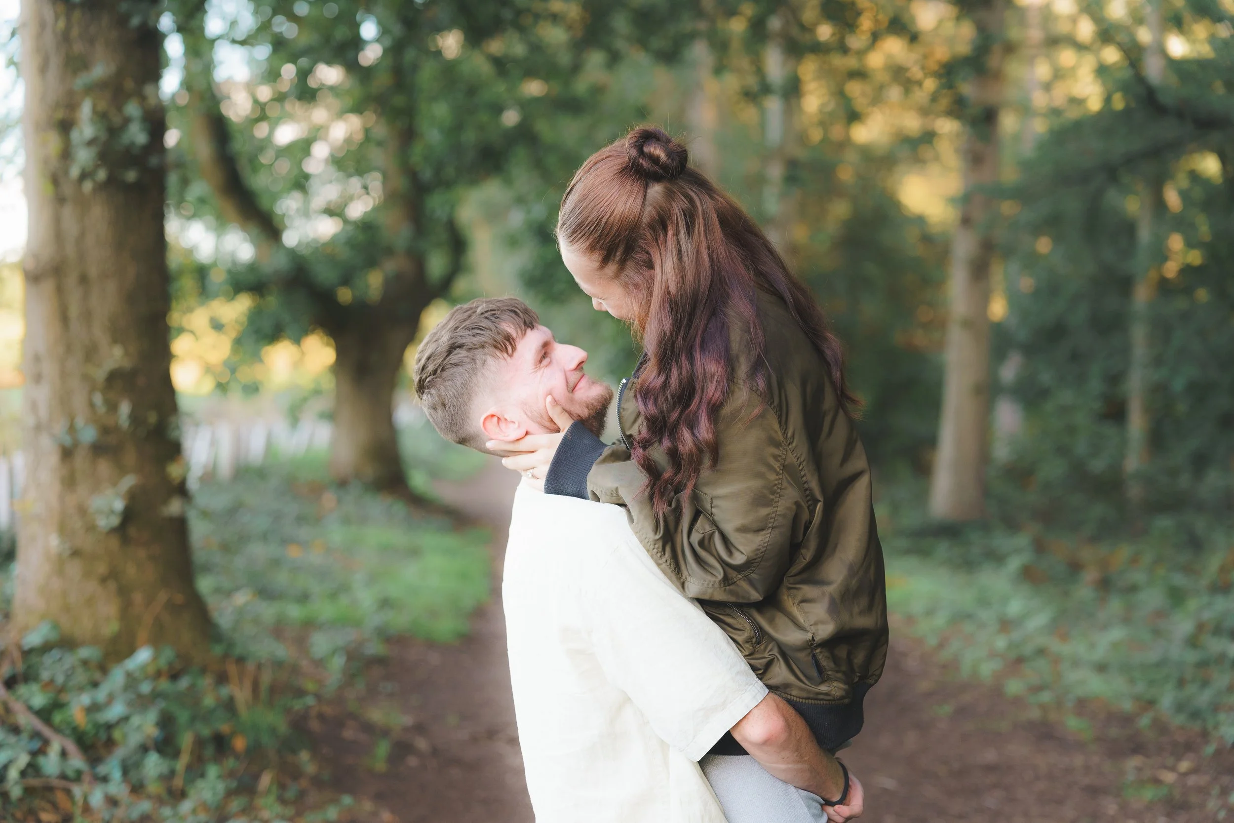 A man holding a woman in his arms in a park with green trees in the background, both smiling and looking at each other.