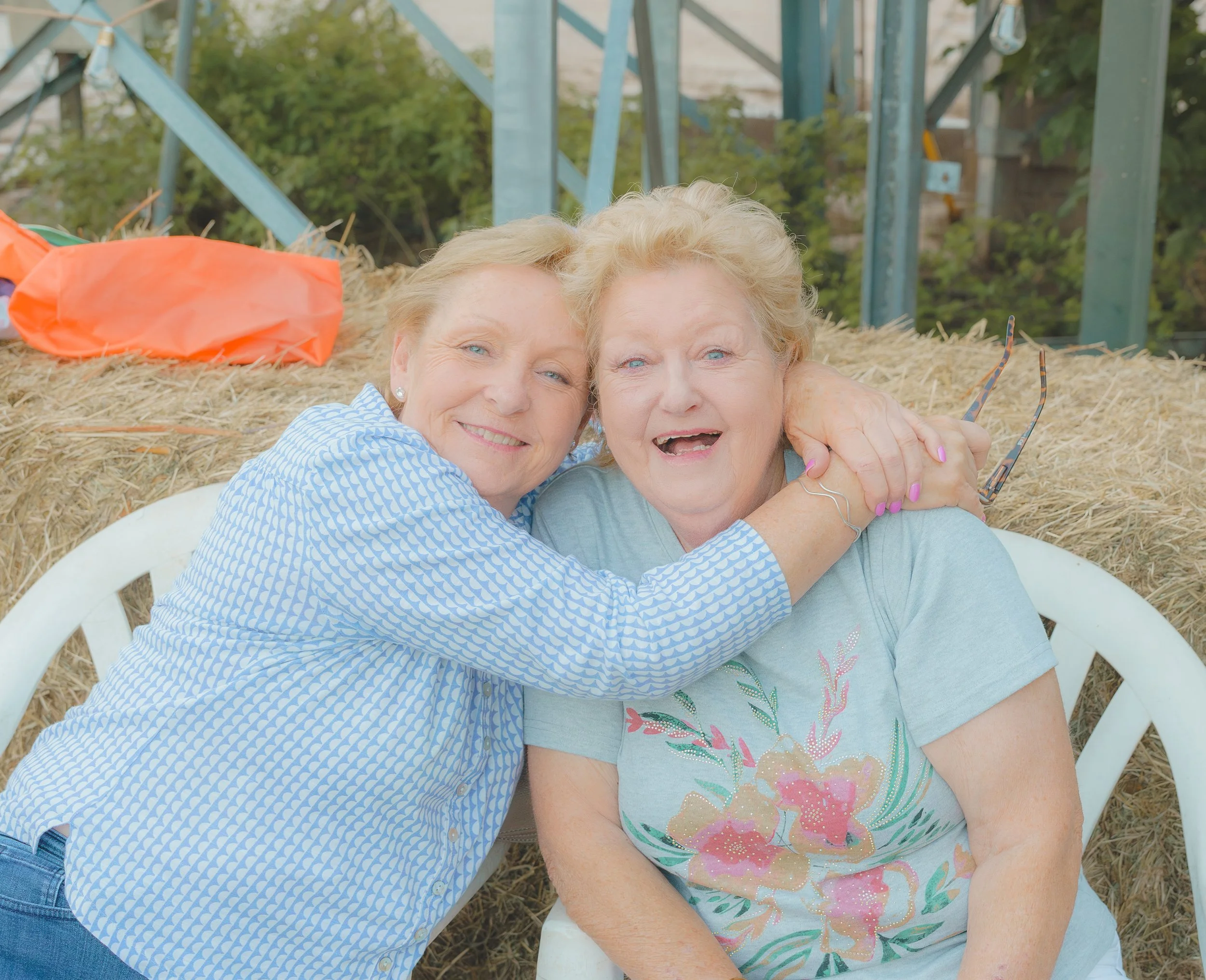 Two elderly women hugging and smiling outdoors on hay surrounded by trees and farm equipment.