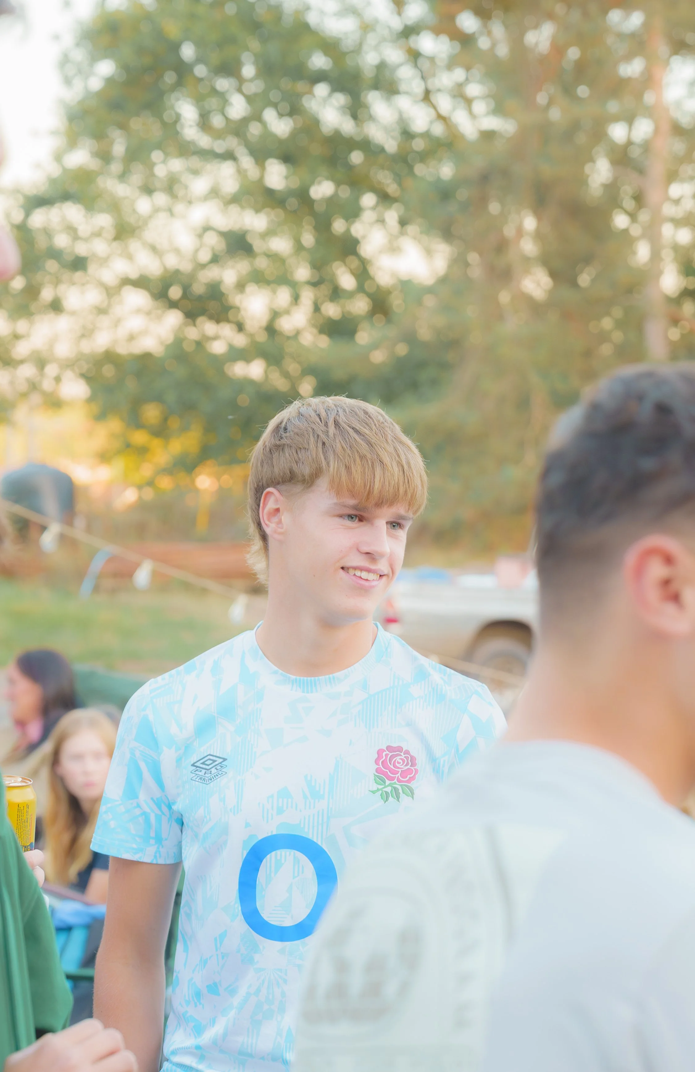 A young man with short, light brown hair, smiling, wearing a light blue and white soccer jersey, standing outdoors among other people, with trees and a blurred crowd in the background.