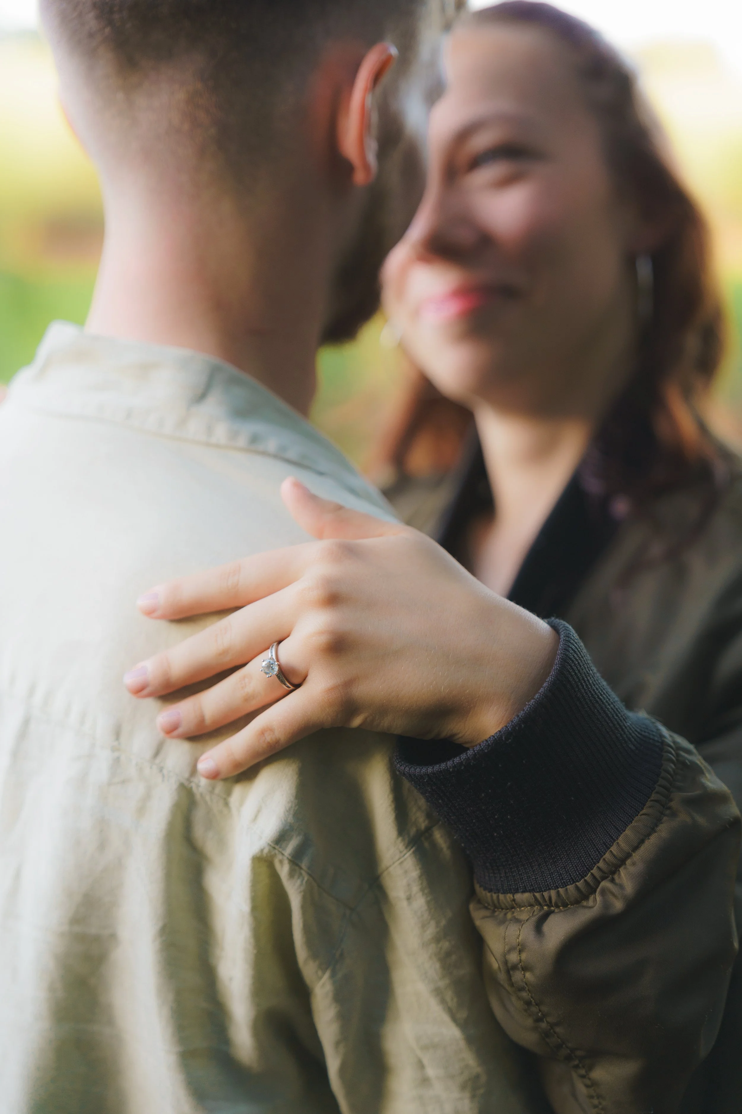 Close-up of a couple embracing, focusing on their faces and hands. The woman is wearing an engagement ring. The background is blurred greenery.