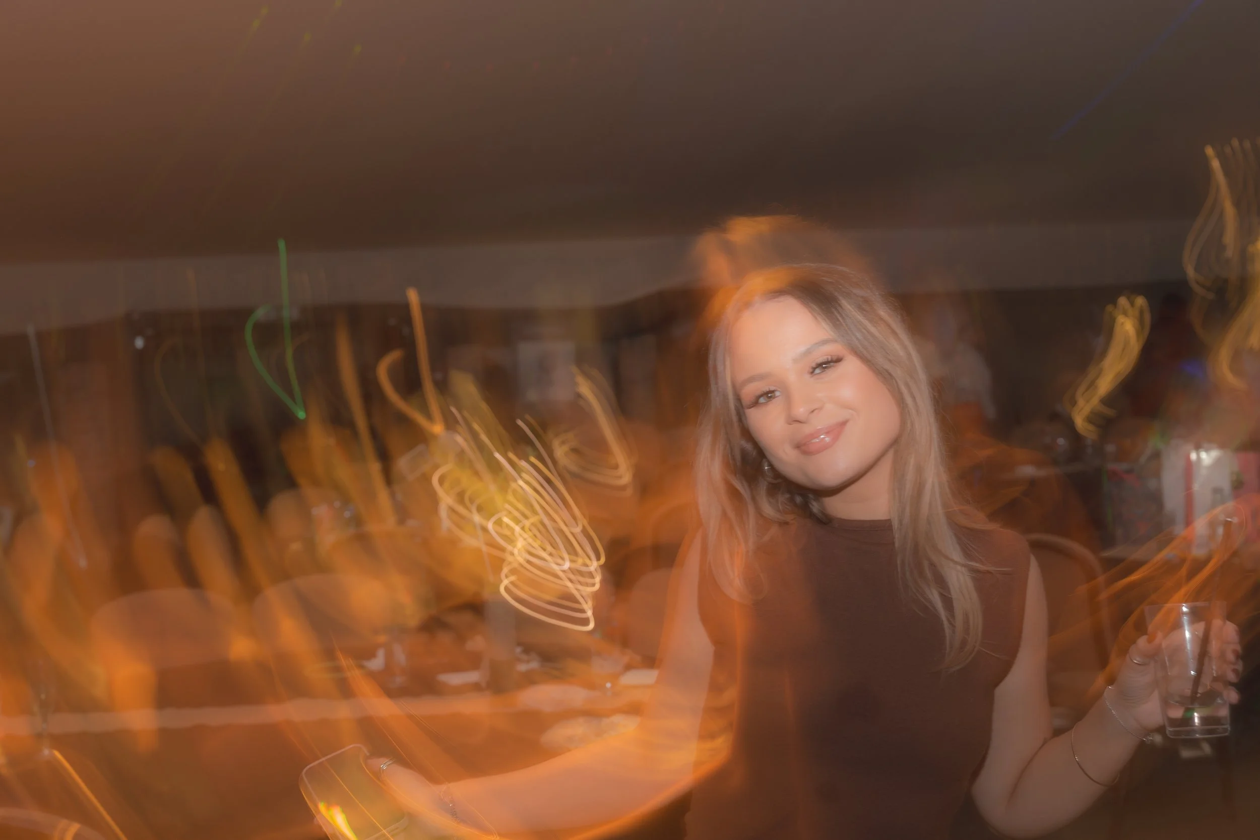 A young woman with long blonde hair smiling and holding a drink in a glass, with colorful light trails in the background.